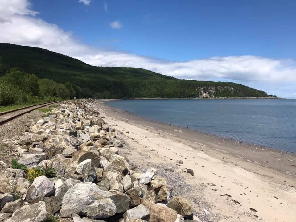 Littoral rocheux avec une plage de sable, voie ferrée et collines verdoyantes sous un ciel bleu.