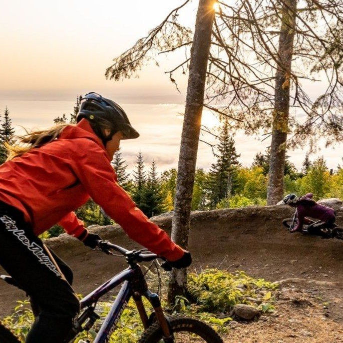 Deux vététistes roulent sur un sentier de terre en forêt, éclairés par le soleil couchant.