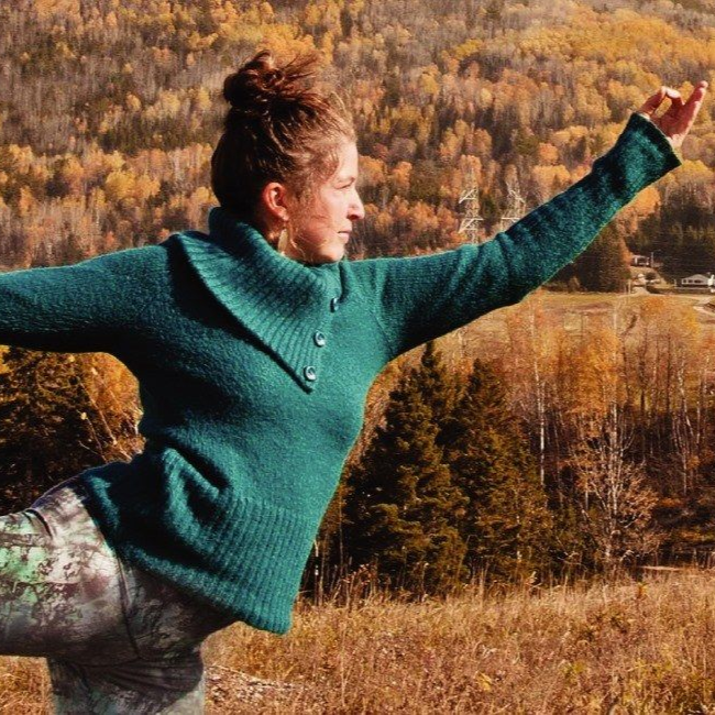 Woman in green sweater balances in a yoga pose outdoors with autumn trees in the background.