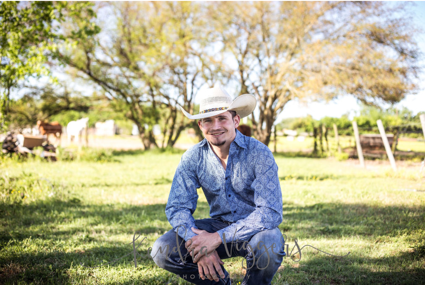 Cowboy in a blue shirt and hat squatting in a field, trees and horses in the background.
