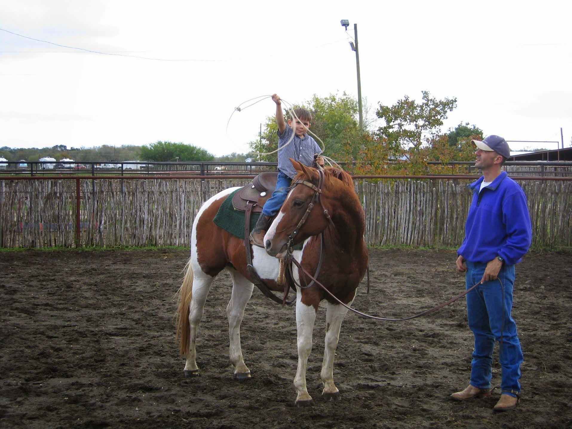 Boy on painted horse practicing lasso, trainer watching in a dirt arena.
