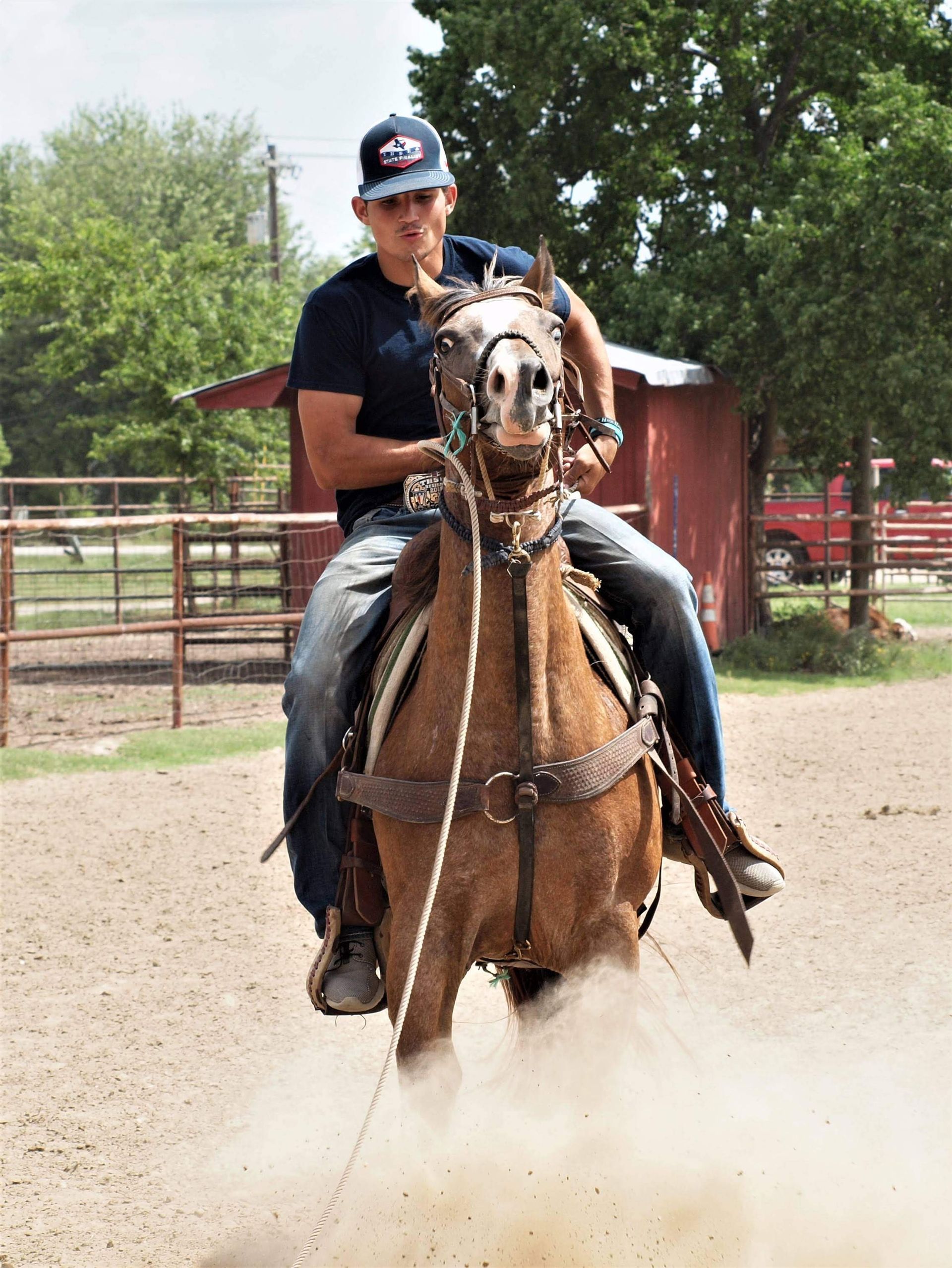 Man riding a brown horse in a dusty arena, wearing a hat and jeans, red barn in the background.