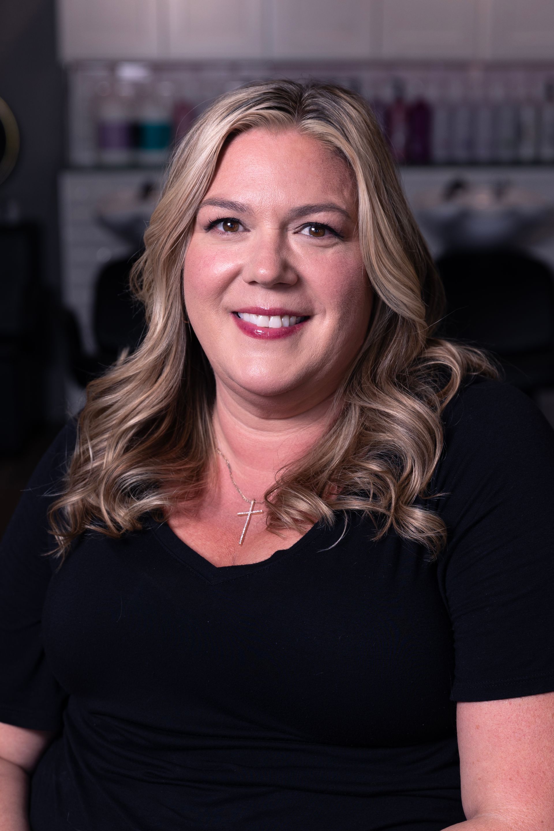 Woman with blonde wavy hair smiles in a salon setting, wearing a black top.