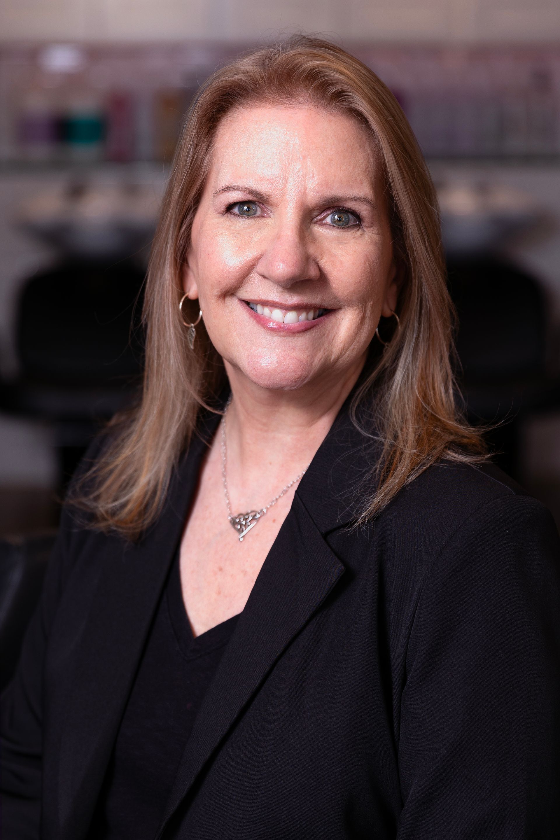 Woman with blonde hair wearing black blazer, smiling in front of a blurred background.