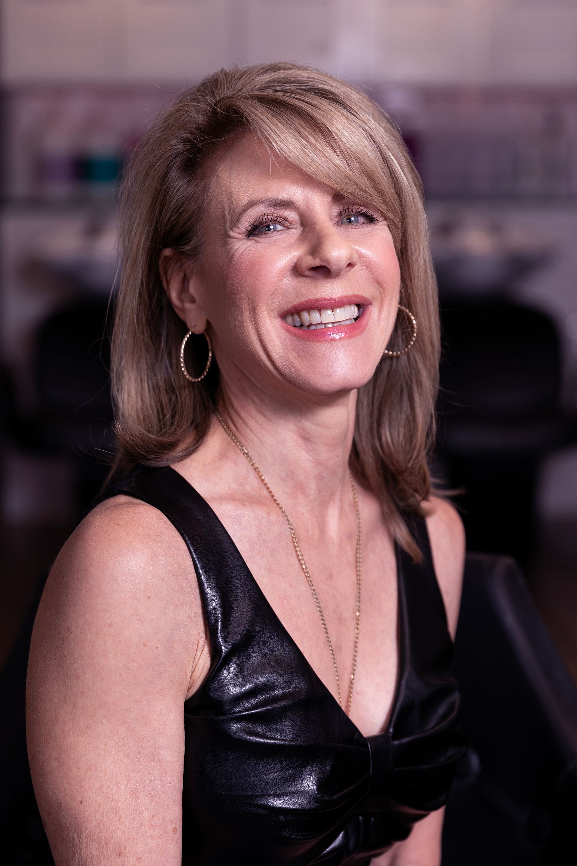 Woman smiling, blonde hair, wearing a black sleeveless top, gold jewelry, and a salon background.