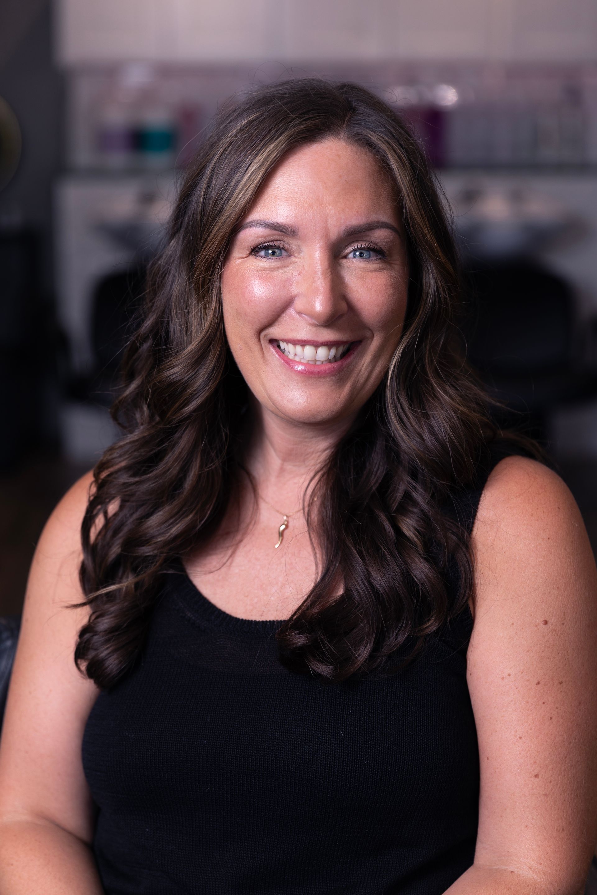 Woman with brown hair smiles, wearing a black top, in a salon.