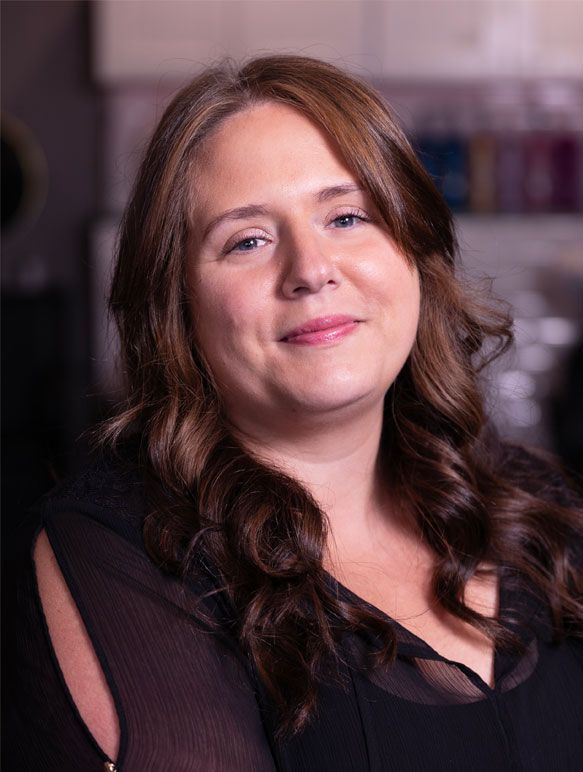 Woman with long wavy brown hair smiles, wearing a black top. Blurred background.