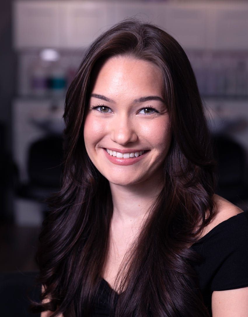 Woman with long, dark hair smiles. She is in a salon and wears a black top.