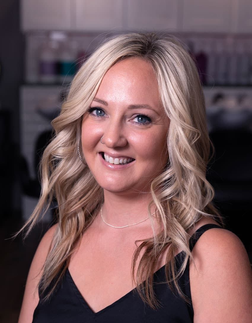 Woman with blonde hair smiling, wearing a black top, in a salon.