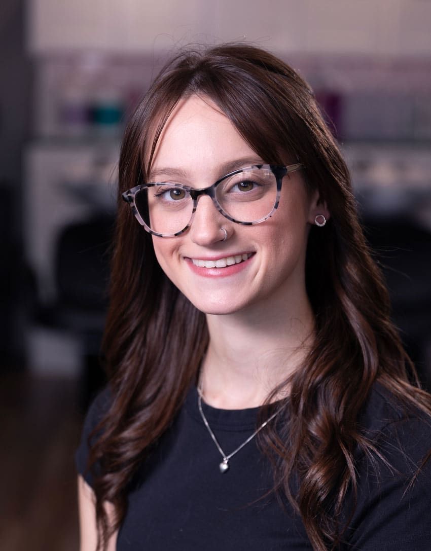 Woman with long brown hair wearing glasses, smiling. In a salon setting, wearing a black shirt.