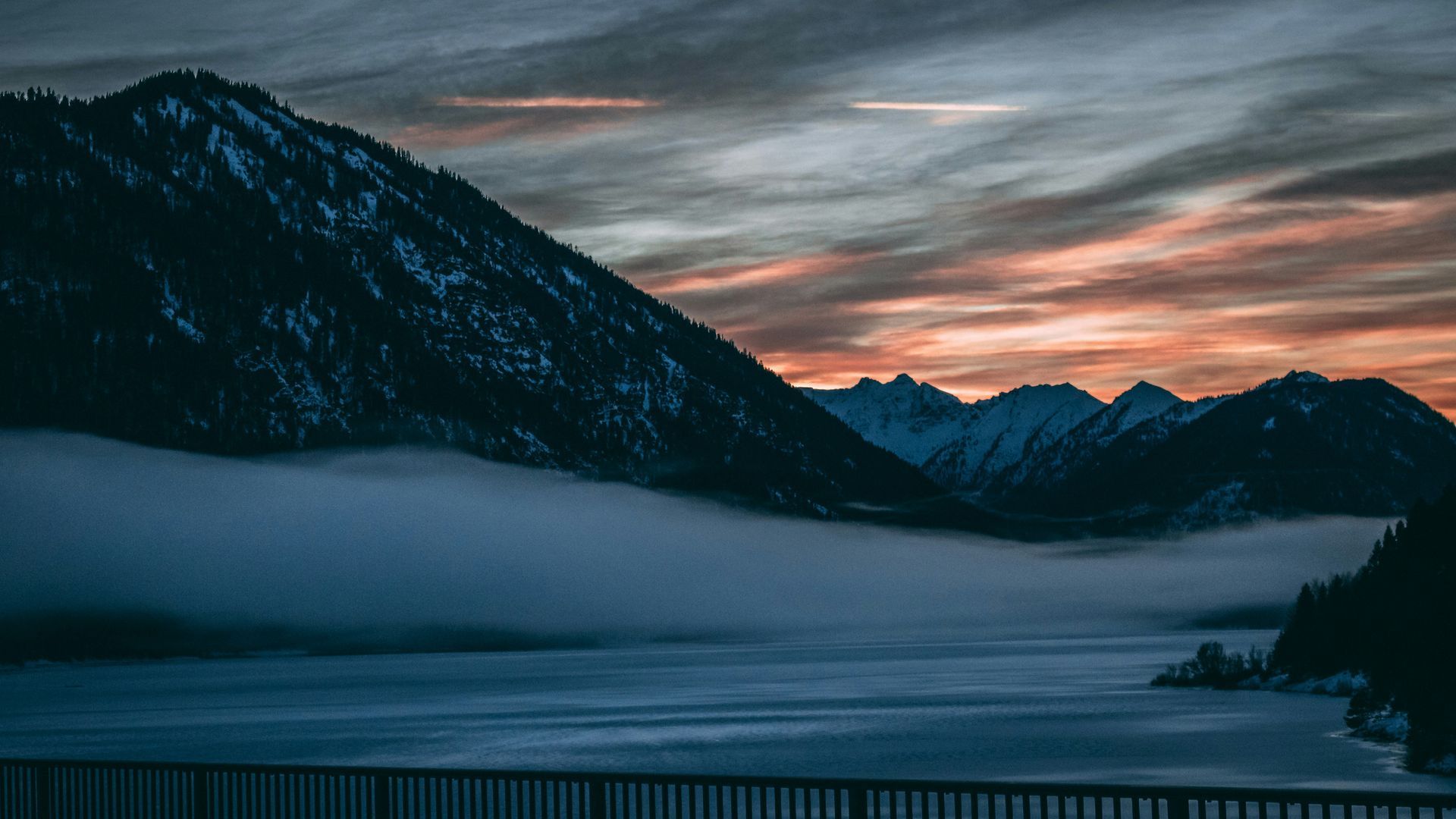 A lake surrounded by snow covered mountains with a sunset in the background.