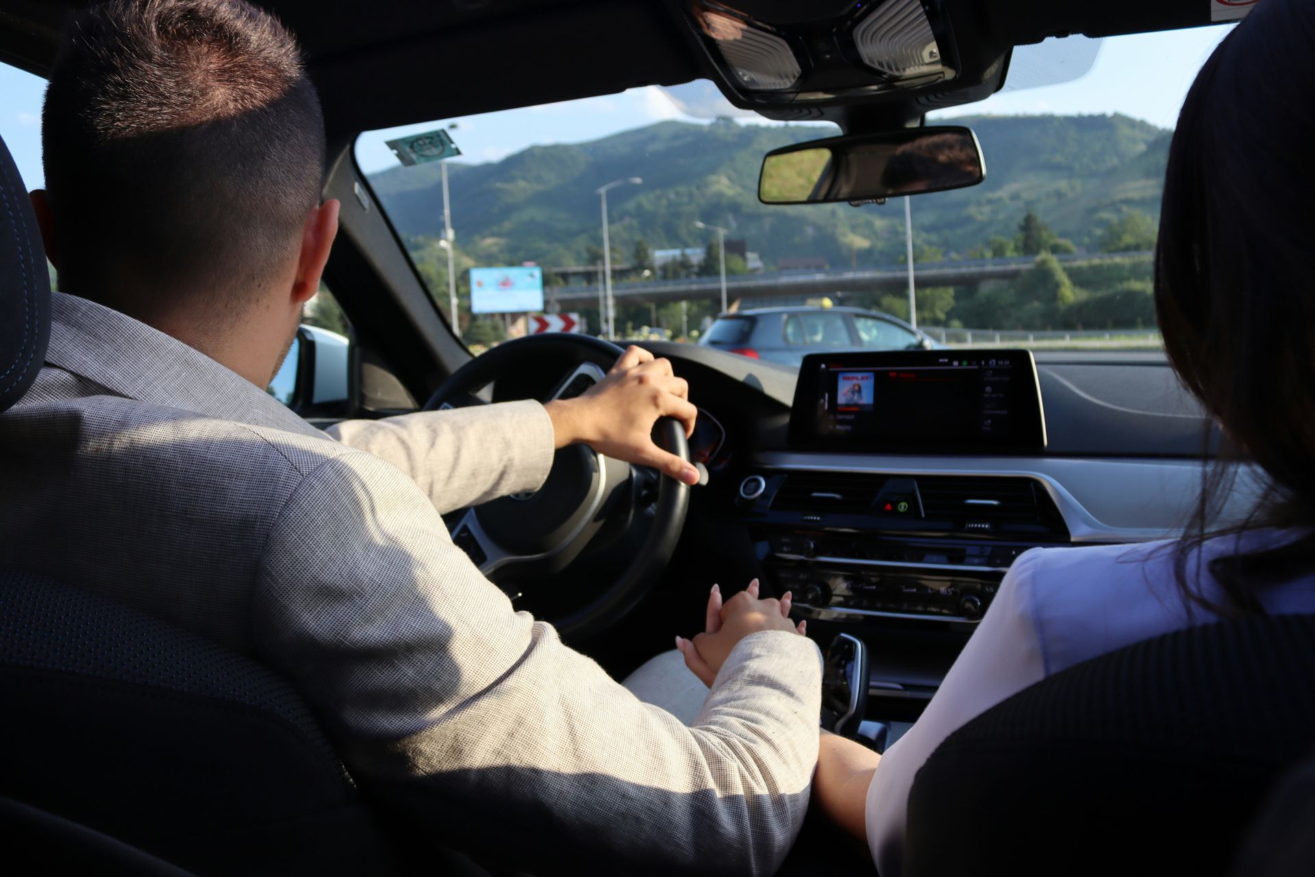 A couple drives on a sunny road, holding hands while the driver steers, with a mountain landscape visible through the glass.