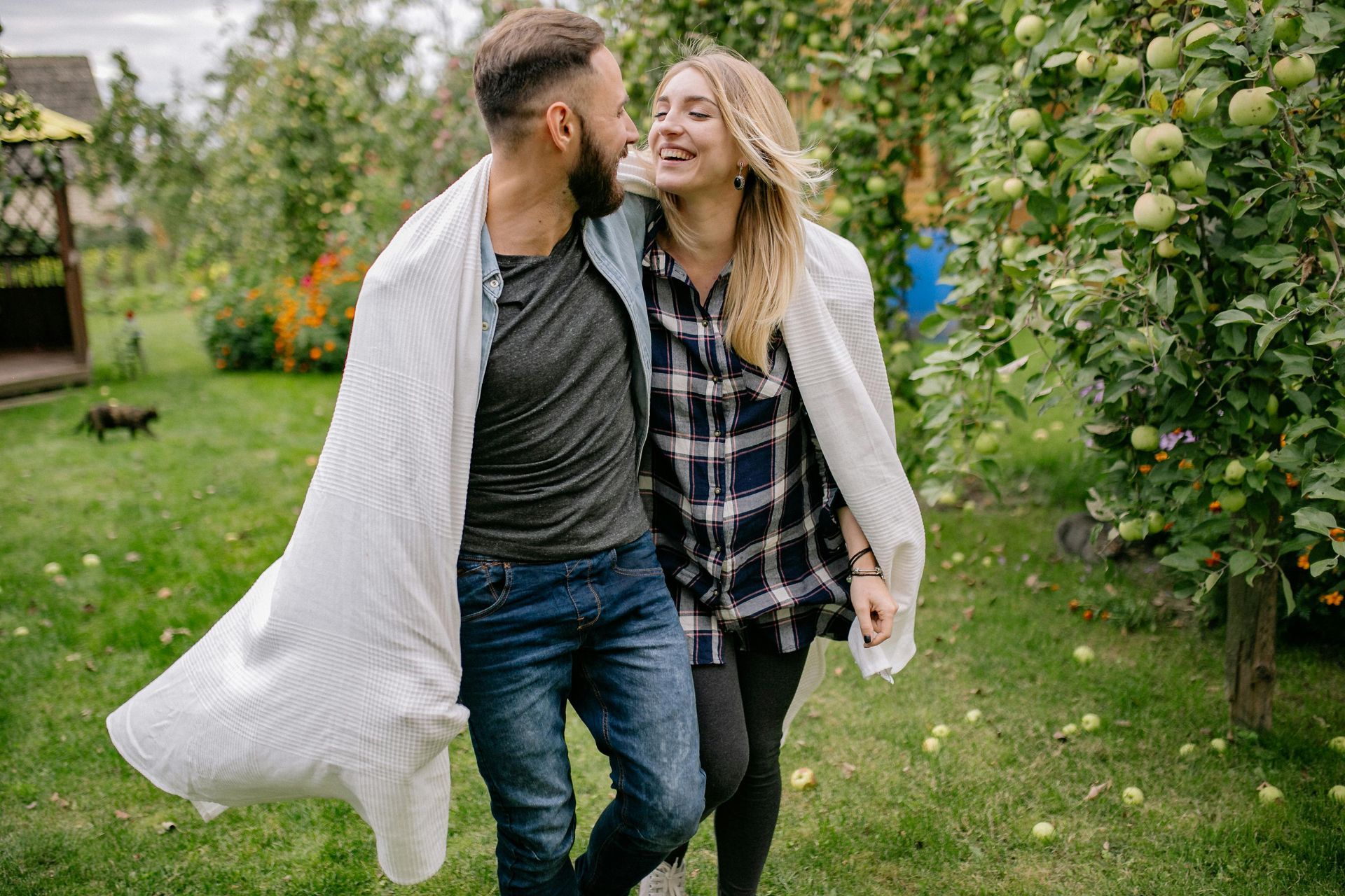 Couple wrapped in white blanket, smiling, walking in a garden.