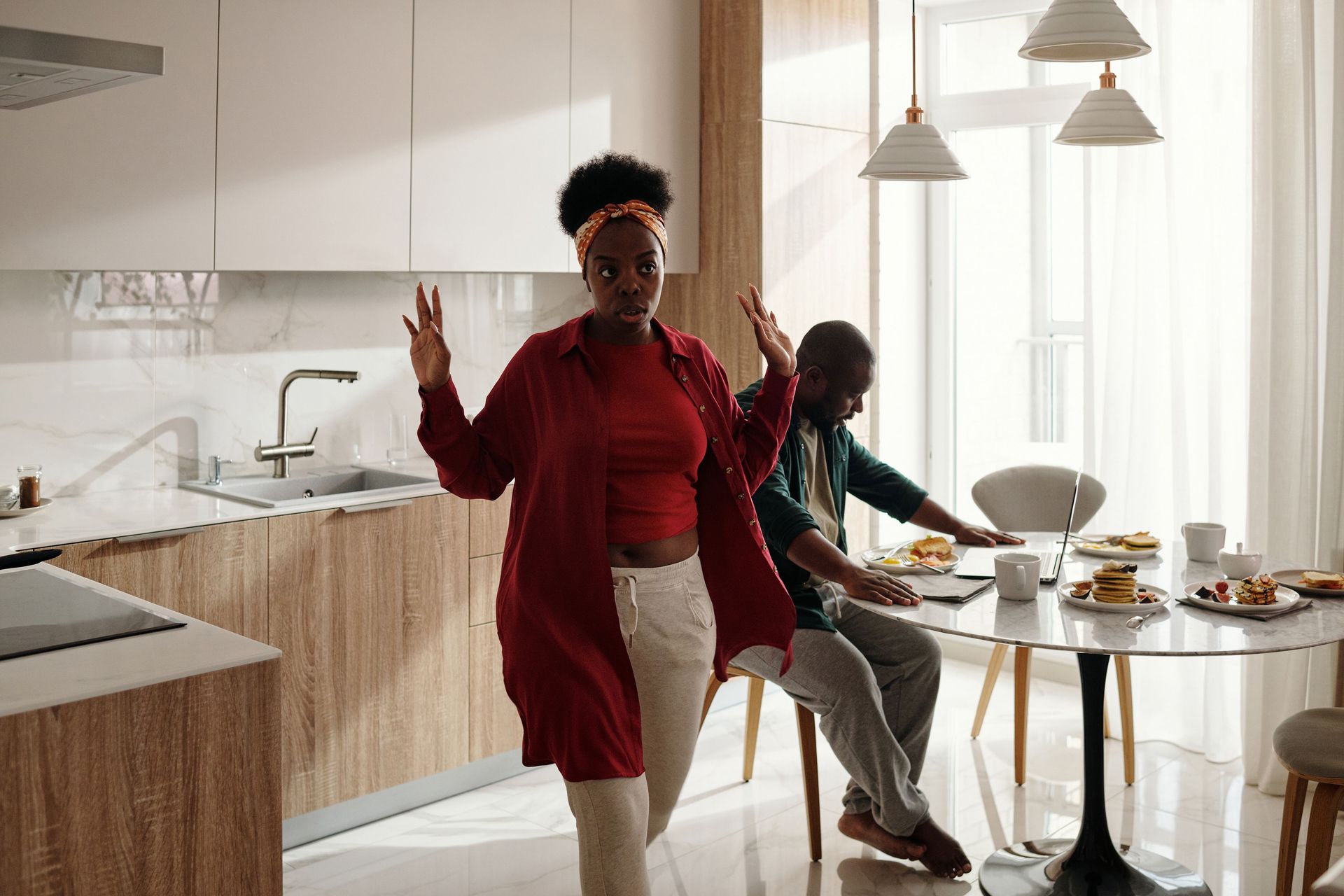 Person dancing in a bright kitchen while another sits at a table with food nearby