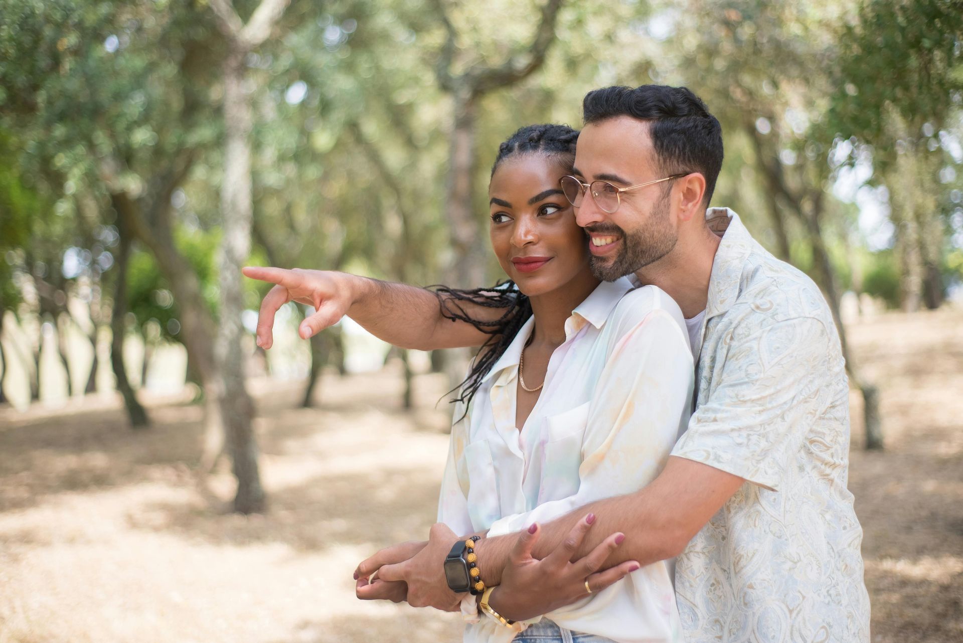 Two people embracing in a sunlit park, smiling as one points into the distance.