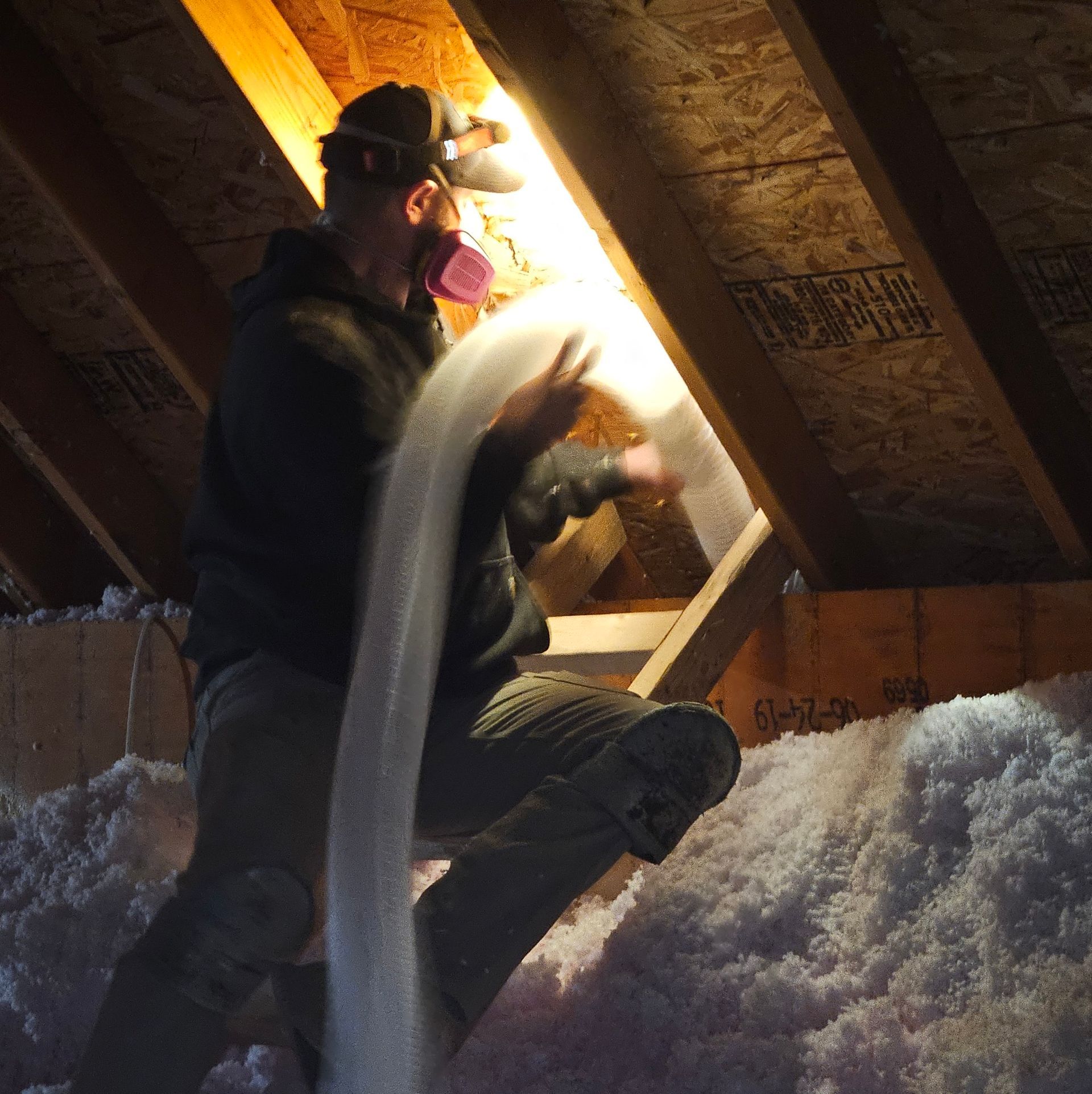 Man in an attic blowing insulation with a respirator.  Bright light shines.