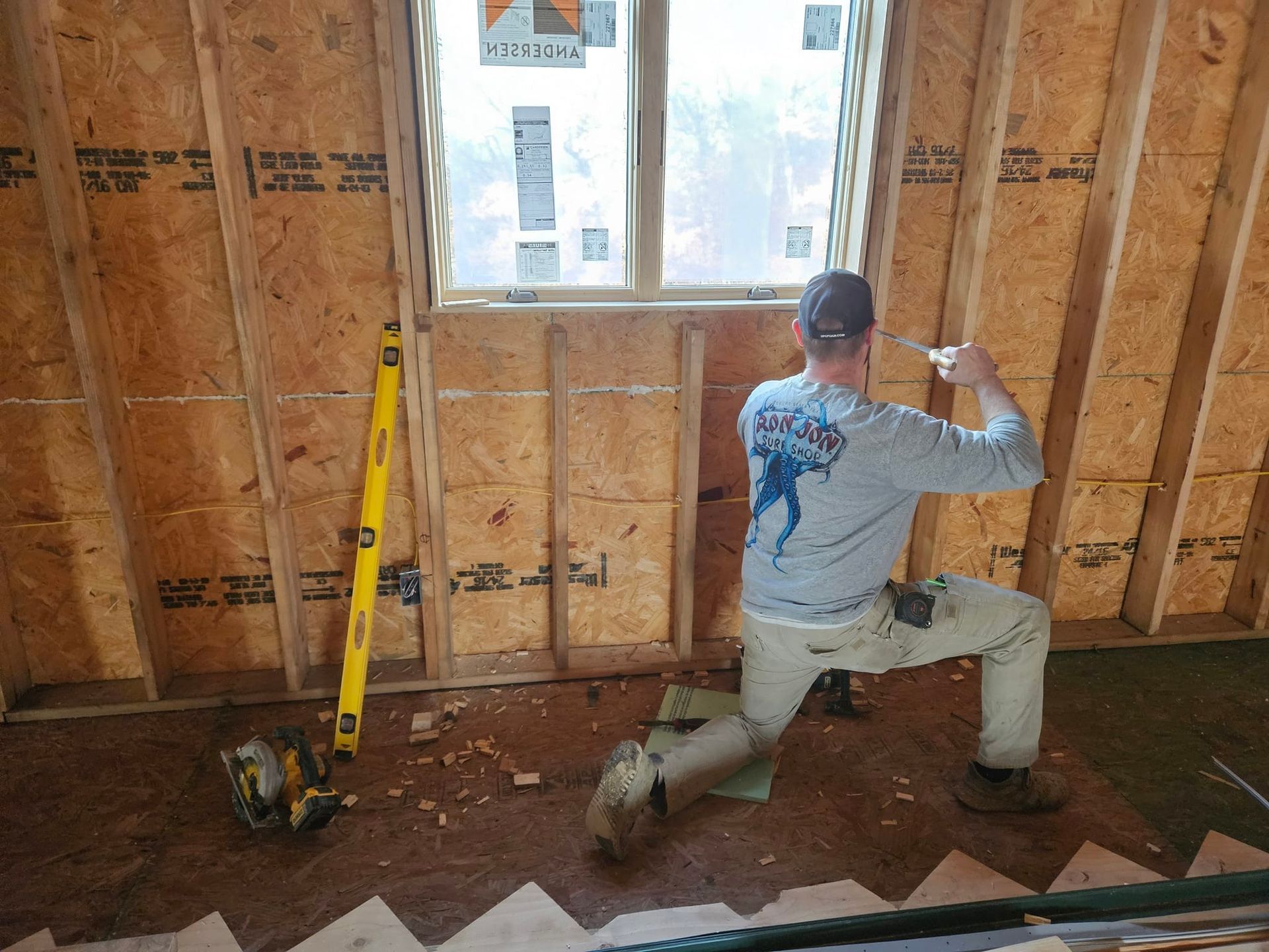 Man installing trim around a window in a wood-framed room under construction.