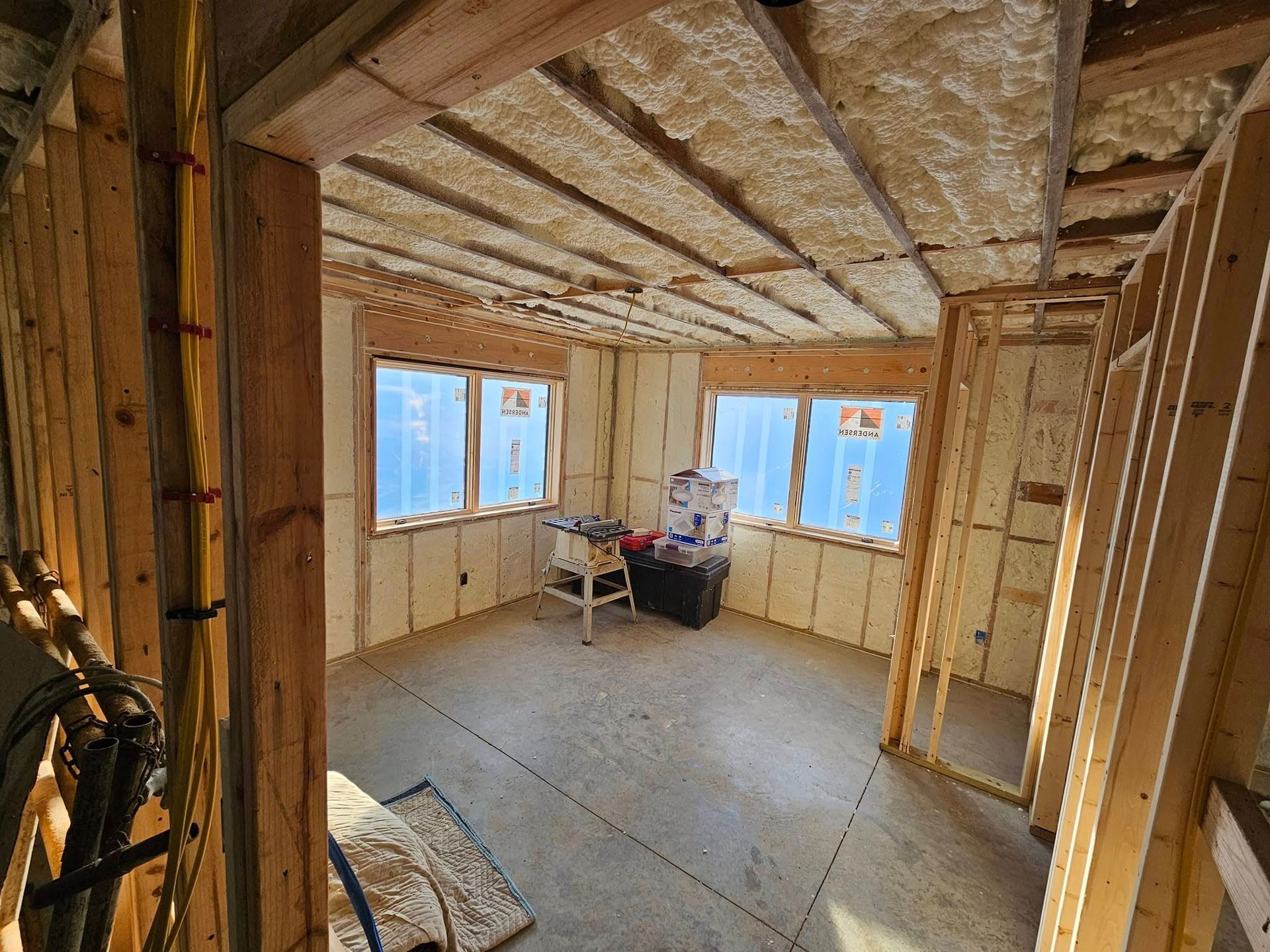 Interior of a room under construction, with exposed wooden framing and spray foam insulation. Two windows visible.