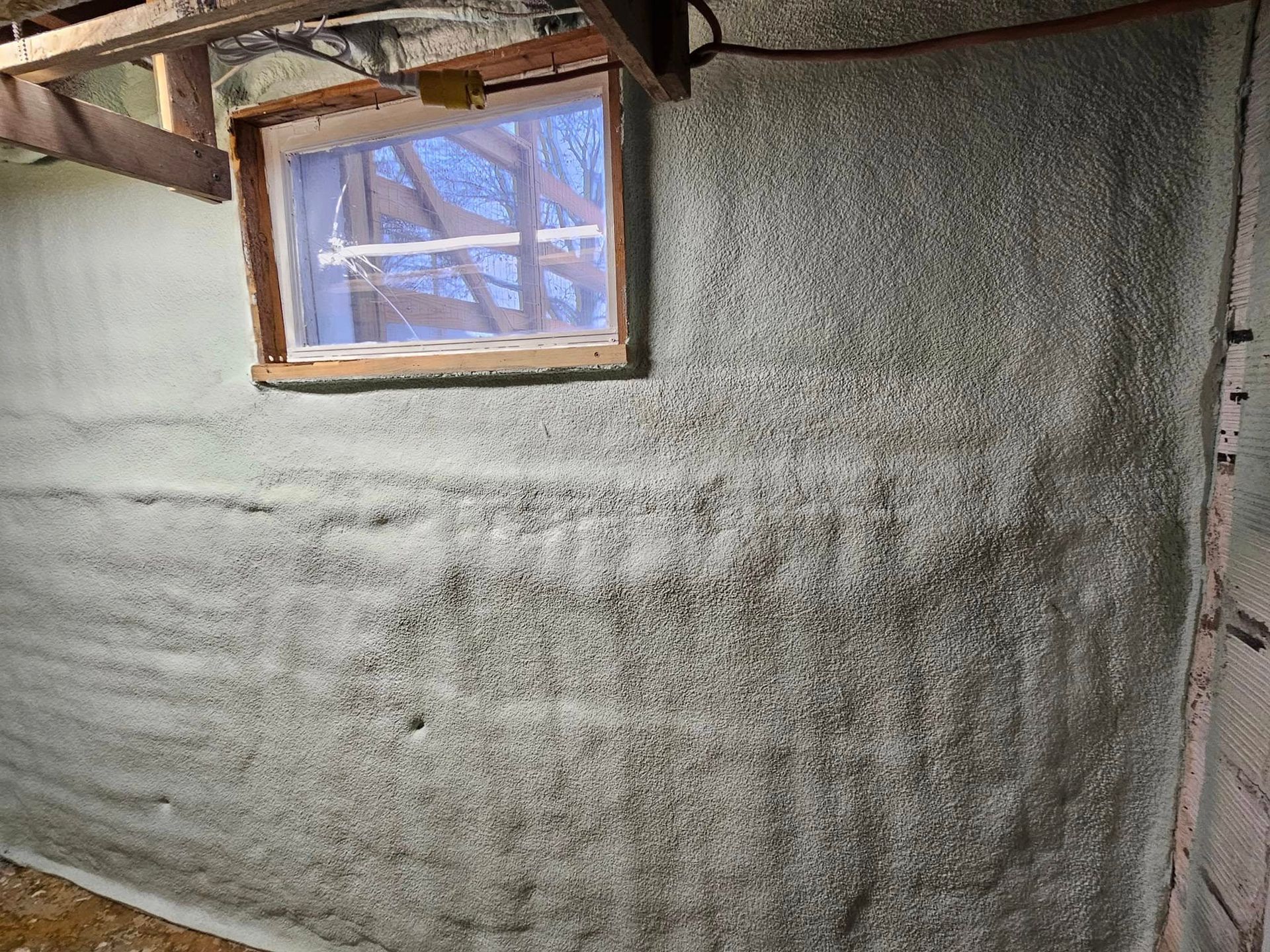 Insulated wall with a small window; spray foam insulation is applied to concrete blocks.
