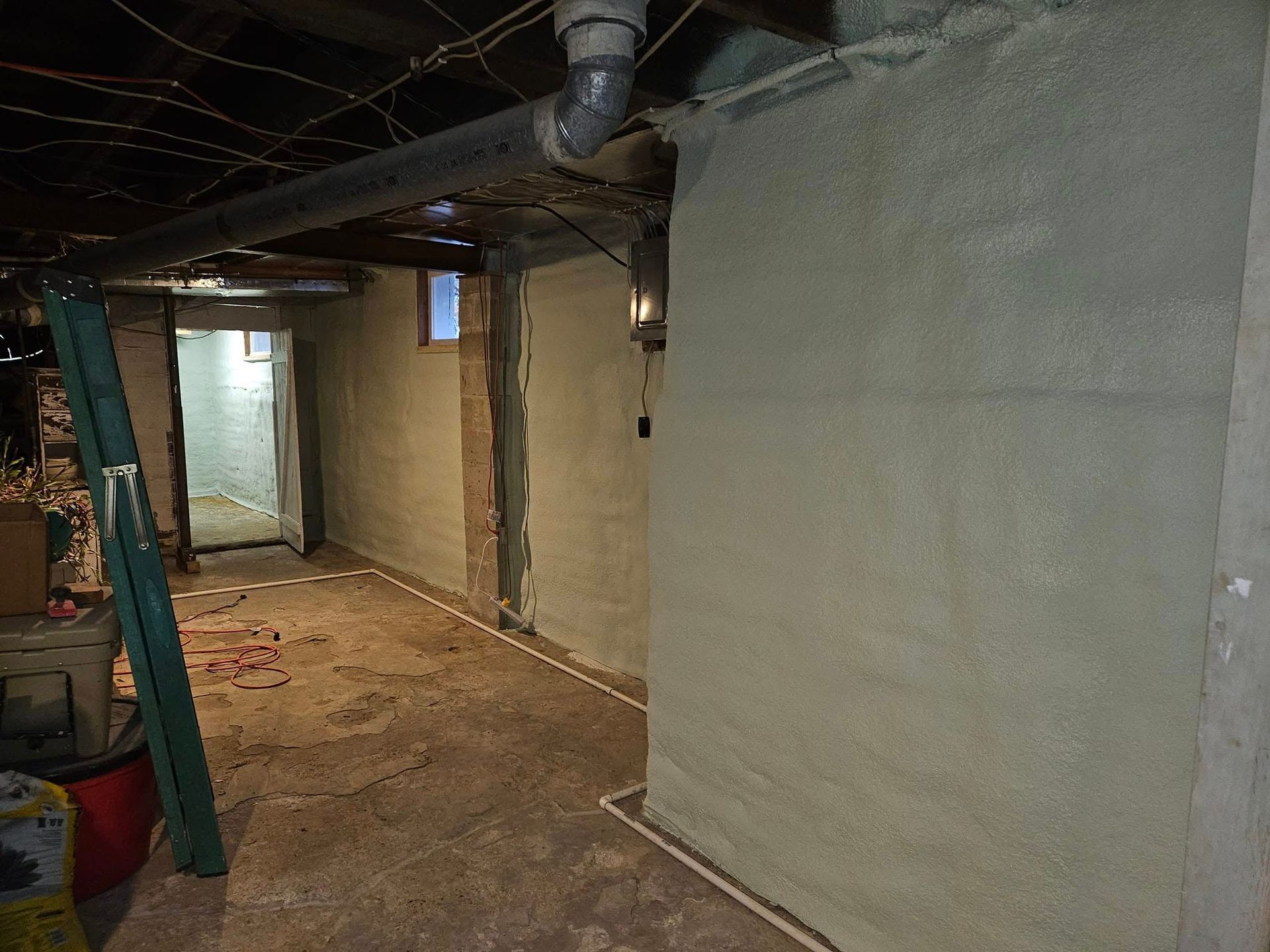 Basement interior with gray walls and concrete floor. Pipes and wiring run overhead. A doorway leads to another room.