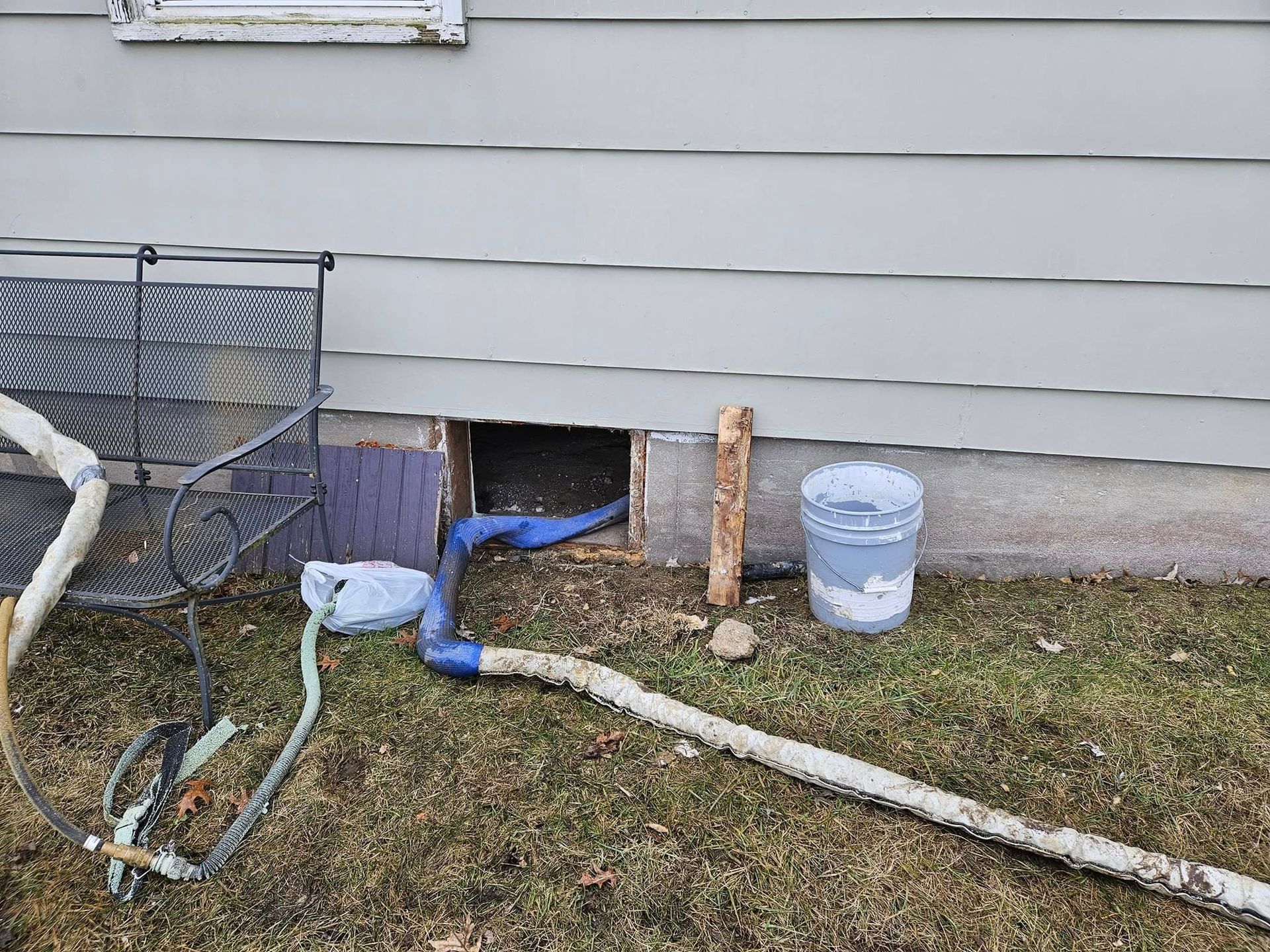 Hole in siding near ground, with blue tubing, a bucket, and a section of wood, likely for work or repair.