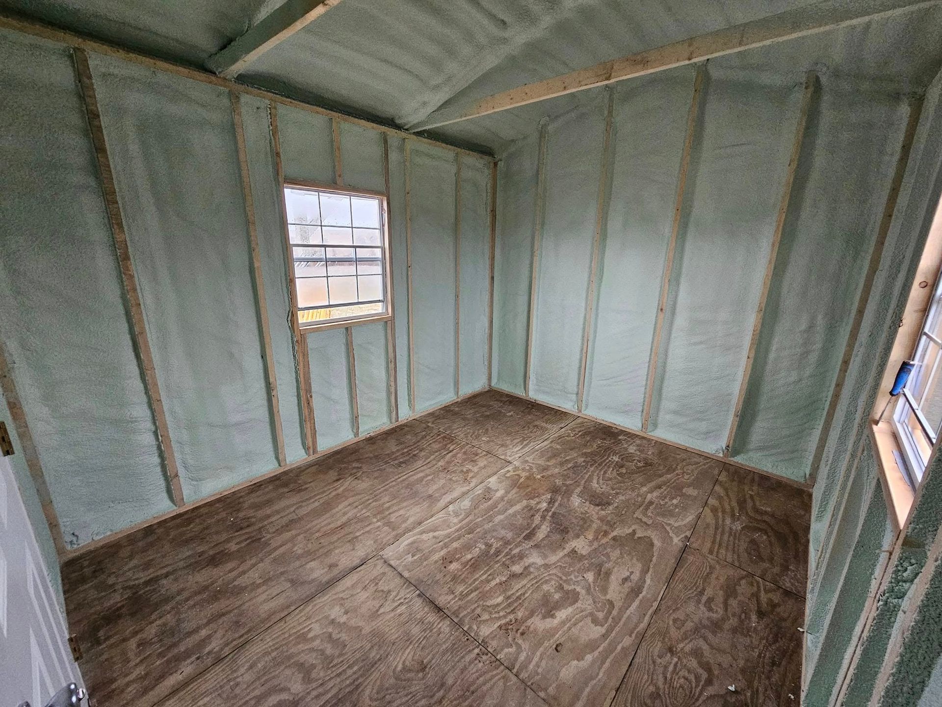 Interior view of a small room with spray foam insulation, a window, and wood flooring.