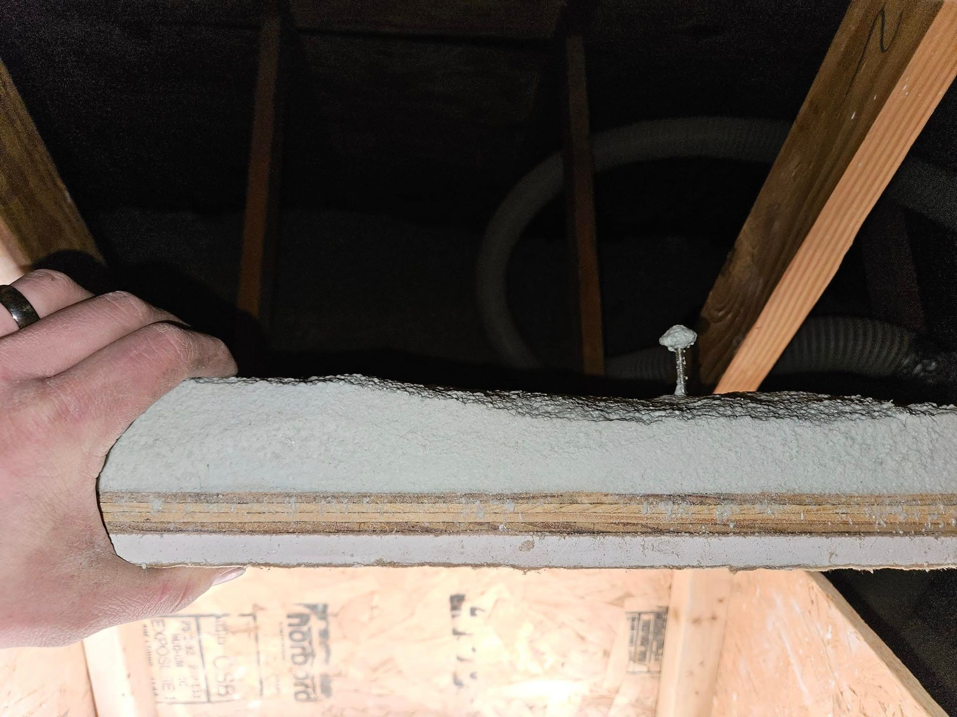 Hand holding a piece of insulation board in an attic. Wood beams and a white vent hose are visible.