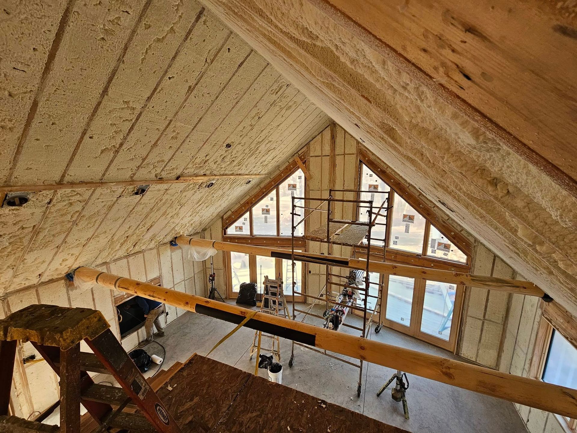 Interior view of a wooden attic with insulation sprayed on the walls and scaffolding in front of windows.