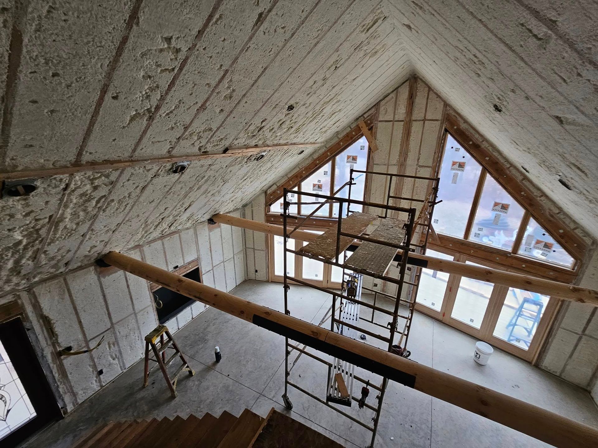 Interior view of a building under construction, spray foam insulation on walls and ceiling and wood beams are visible.