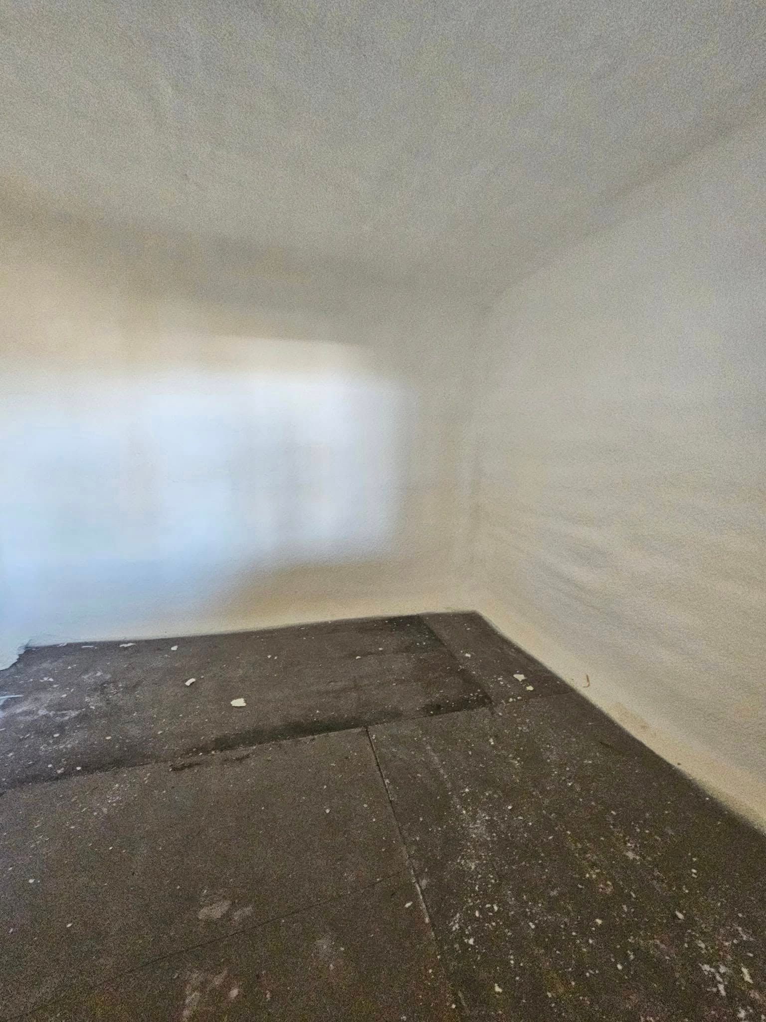 Empty room with dark debris-covered floor and off-white walls. A window creates a bright, blurry light source.