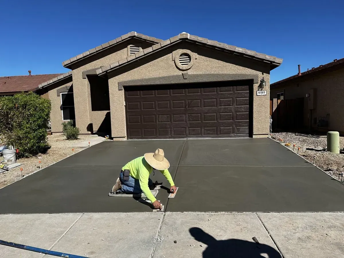 Person smoothing wet concrete driveway in front of a tan house with a dark brown garage door on a sunny day.