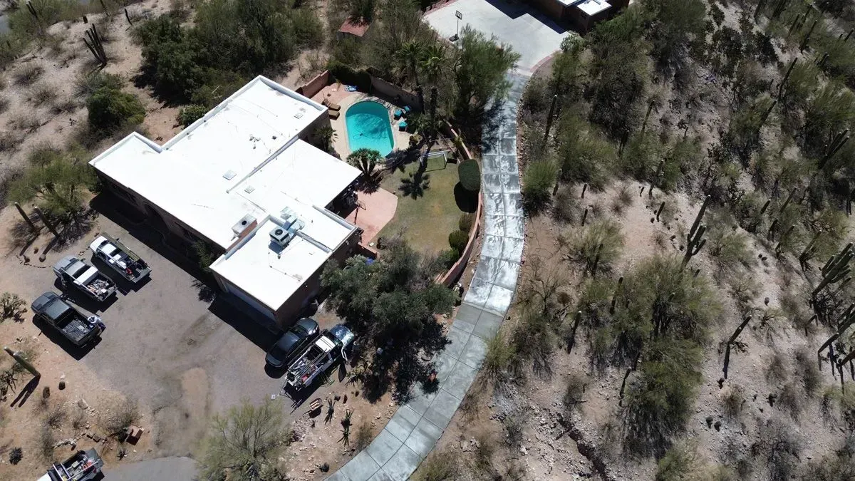 Aerial view of a single-story house with a pool, surrounded by desert vegetation. Several parked vehicles are visible.