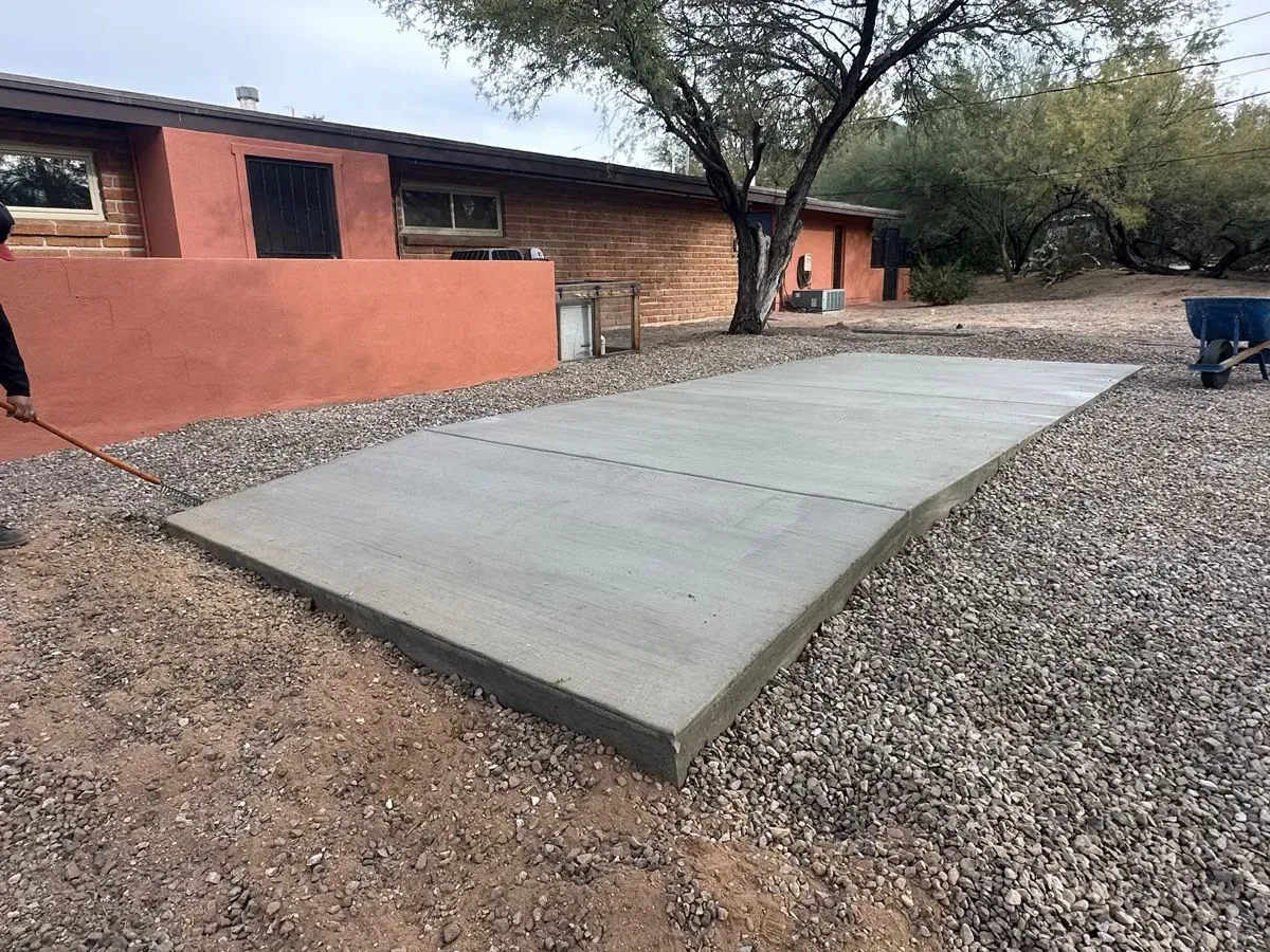 Freshly poured concrete patio with dark gravel surround next to a rustic building.