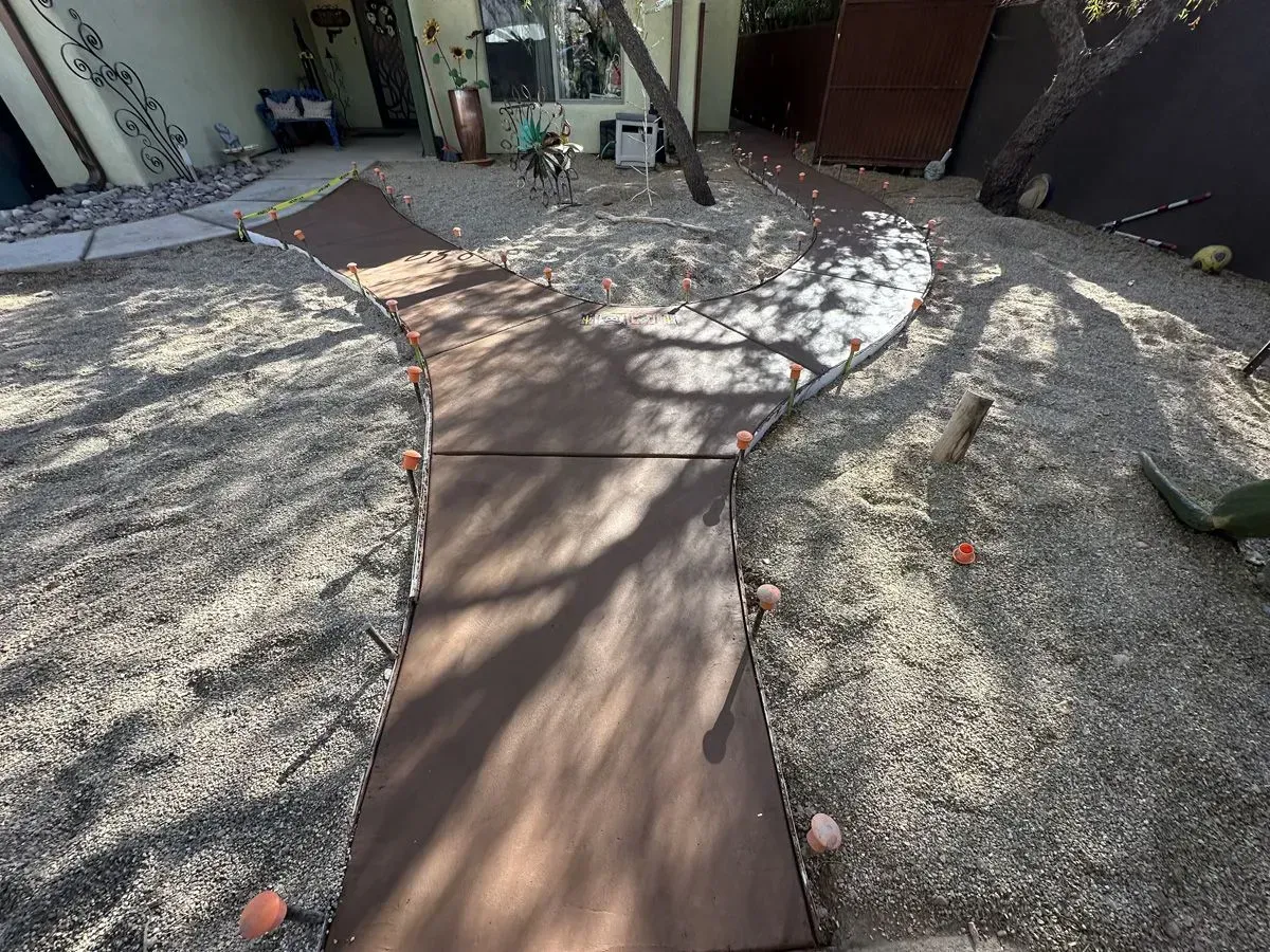 Brown walkway through a gravel yard, leading to a house. Sunlight casts shadows.