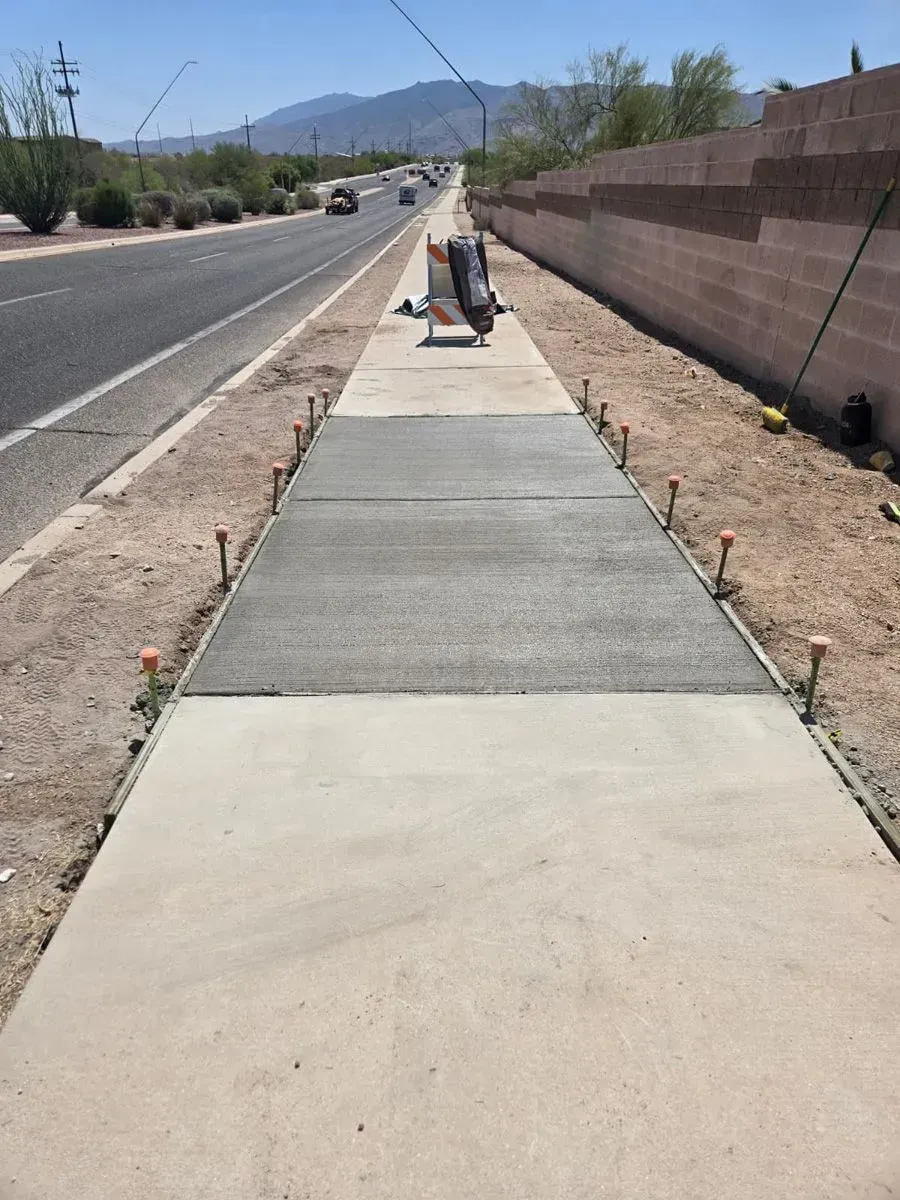 Sidewalk construction: freshly poured concrete between existing sections, with road and mountains in the background.