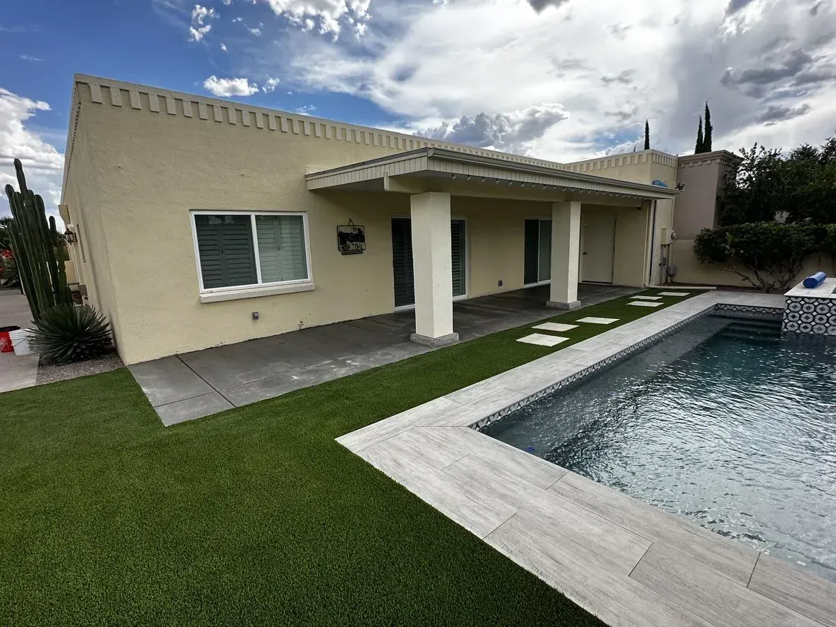 Beige house with a covered patio, swimming pool, and green lawn under a cloudy sky.