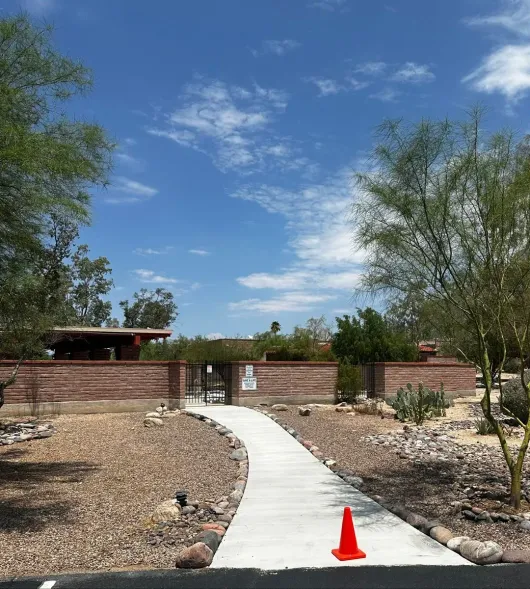 Concrete path leads to a gate within a brick wall, with blue sky and trees. Orange cone in foreground.