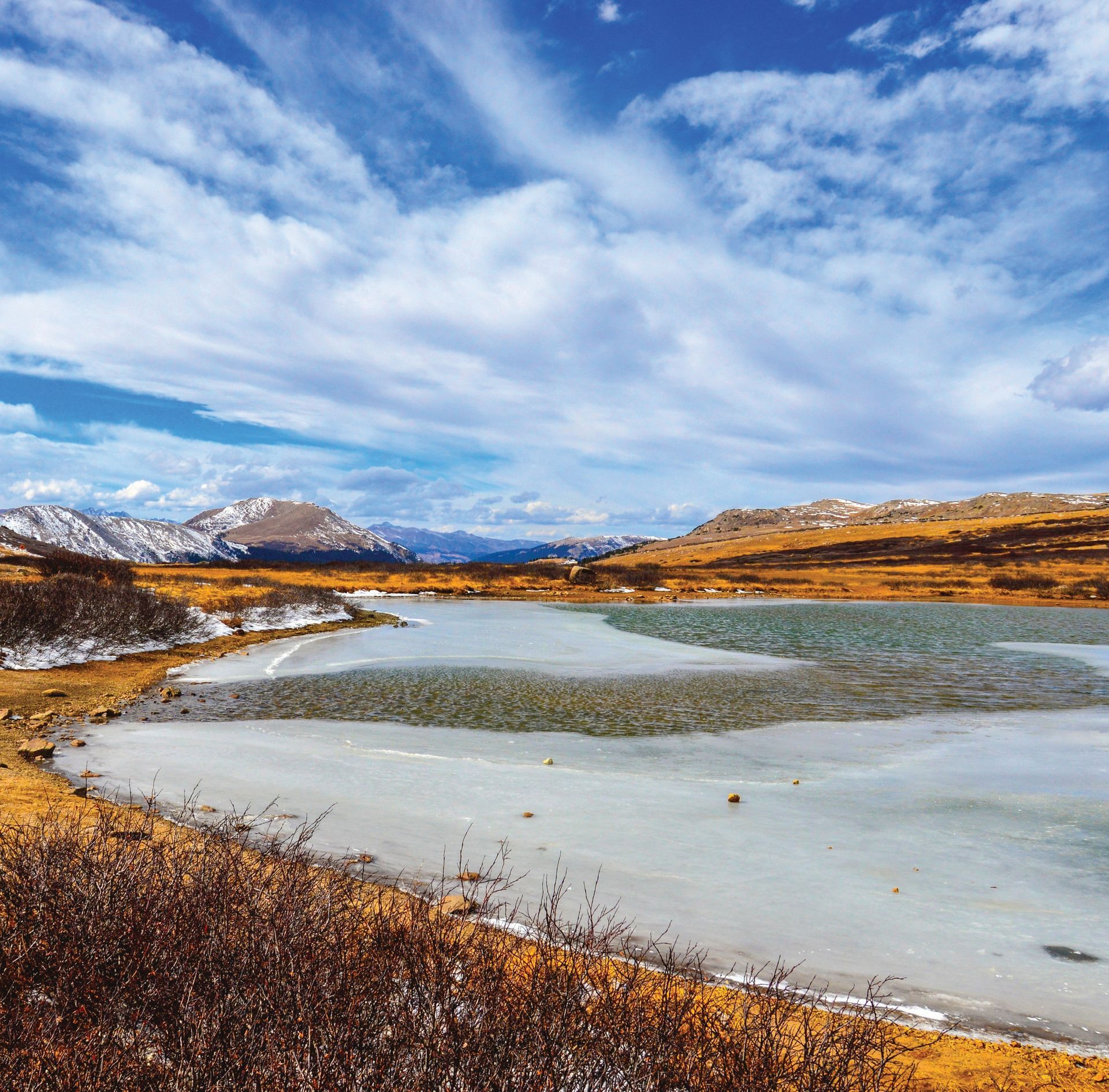 A partly frozen alpine lake set in a golden, grassy landscape under a bright, cloudy blue sky with distant snow-capped hills in colorado