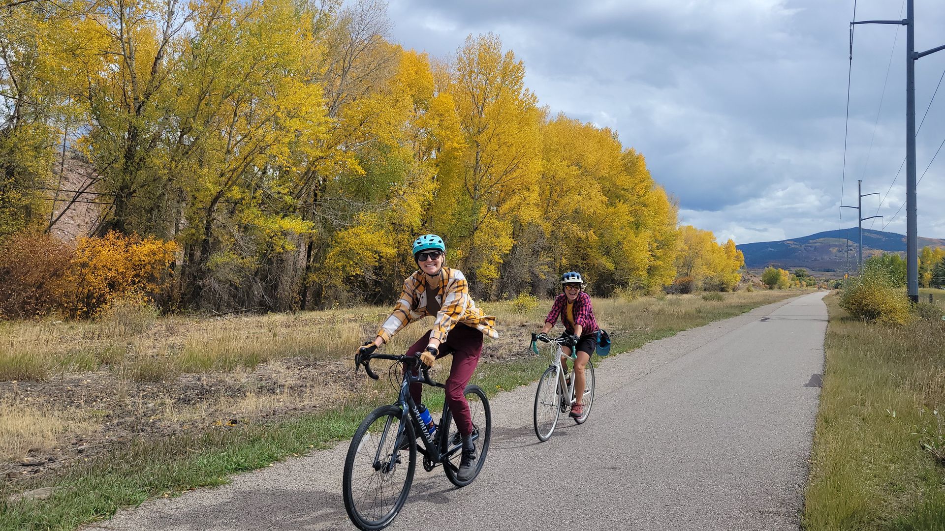Two people cycle along a paved path bordered by autumn trees with bright yellow leaves under a cloudy sky in roaring fork colorado