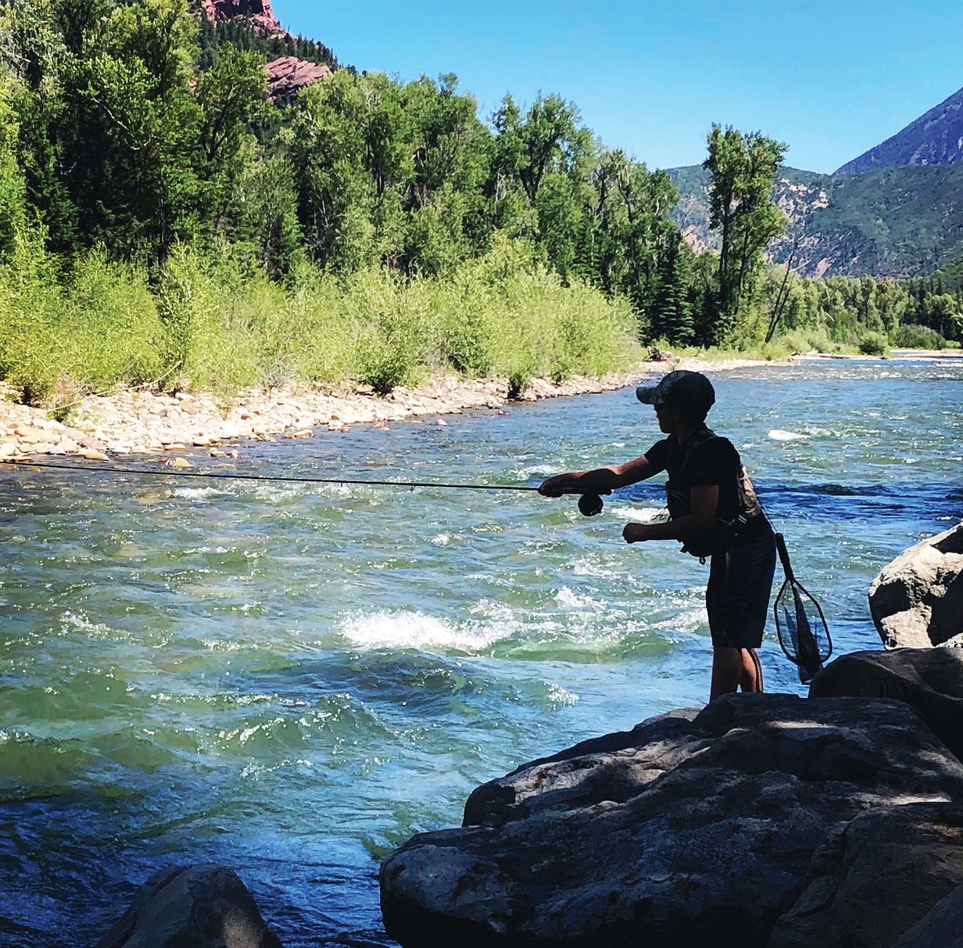 A person fly-fishing in a sunlit river surrounded by trees and mountains.