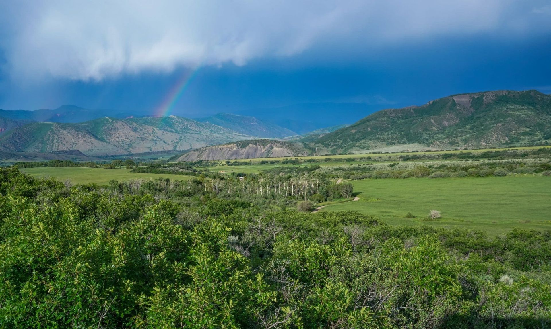A faint rainbow stretches across a dramatic, stormy sky over a green valley bordered by rolling mountains in colorado
