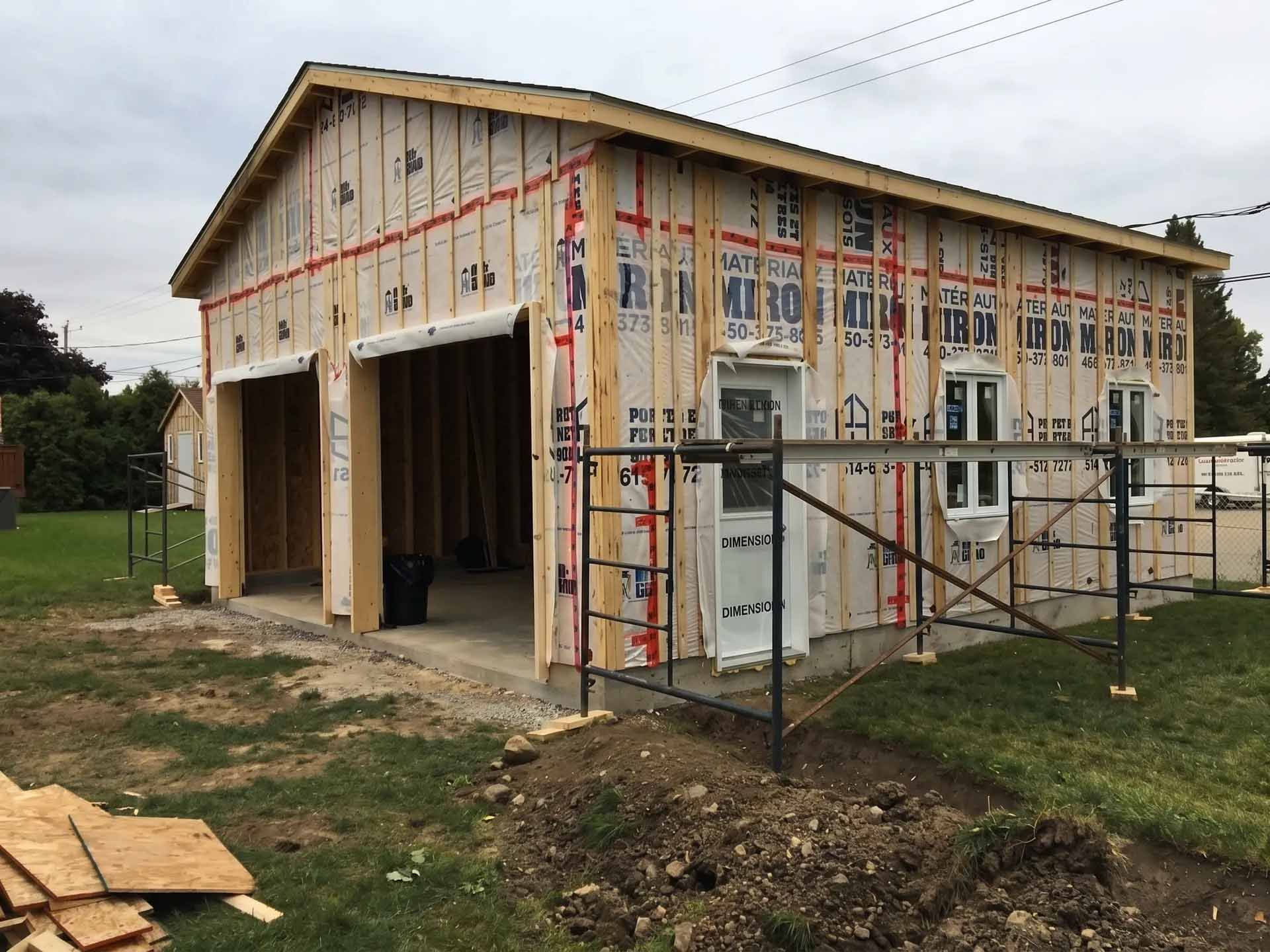 Garage partiellement construit avec isolation apparente, travées ouvertes et échafaudages sur un terrain gazonné