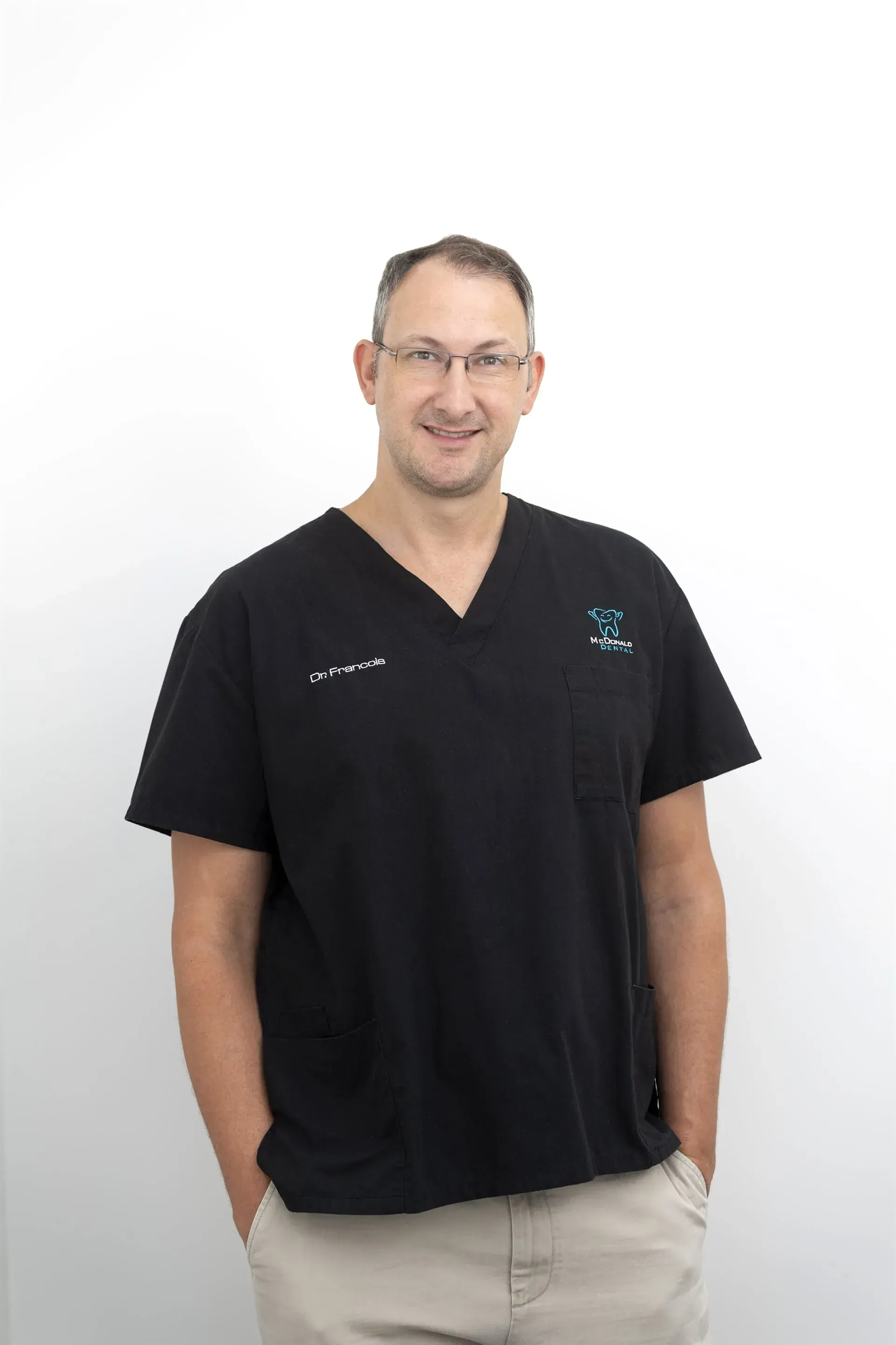 Man in black scrubs and glasses, hands in pockets, smiling. White background. — McDonald Dental in Gympie, QLD