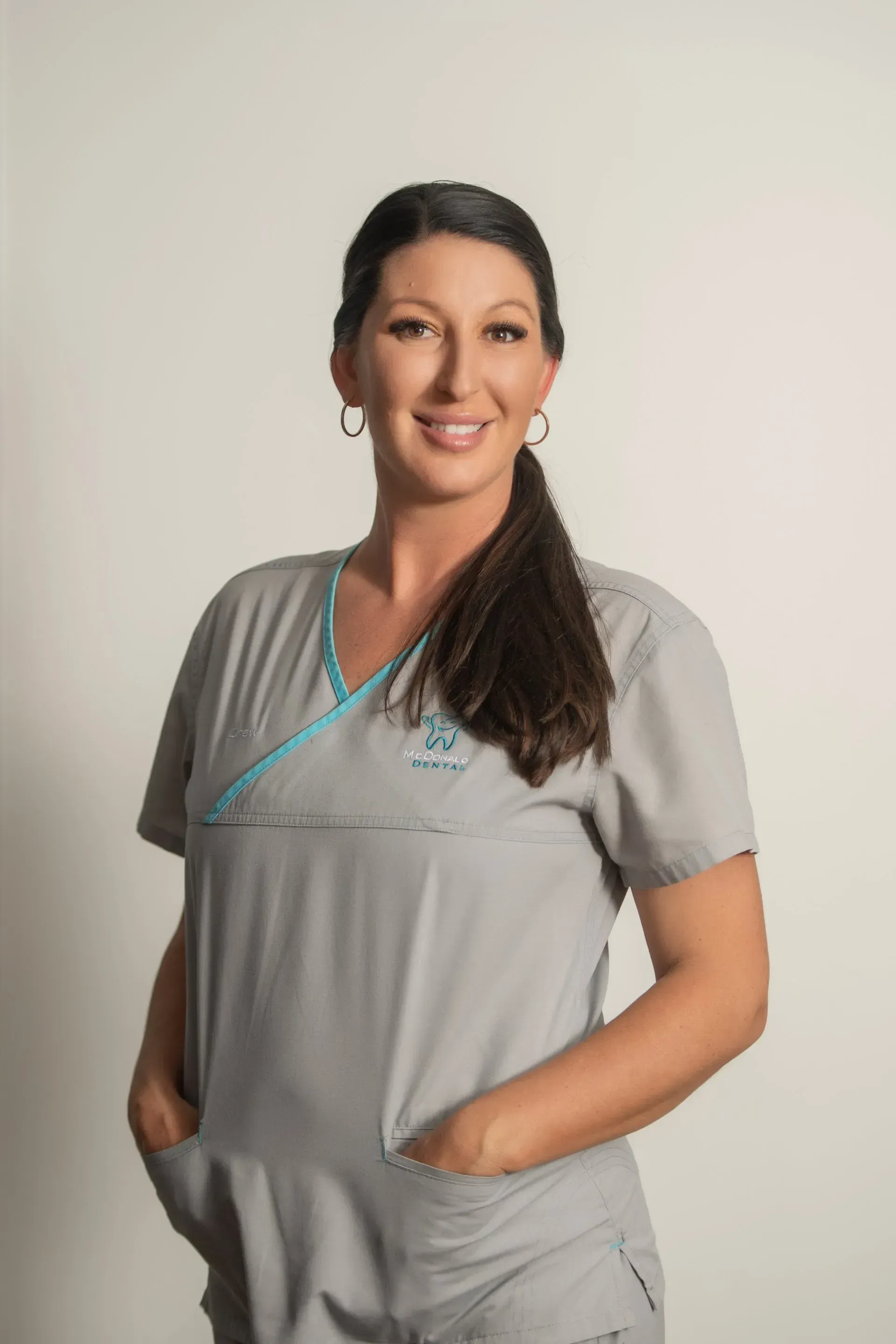 Woman in gray scrubs with teal trim, hands in pockets, smiling. — McDonald Dental in Gympie, QLD