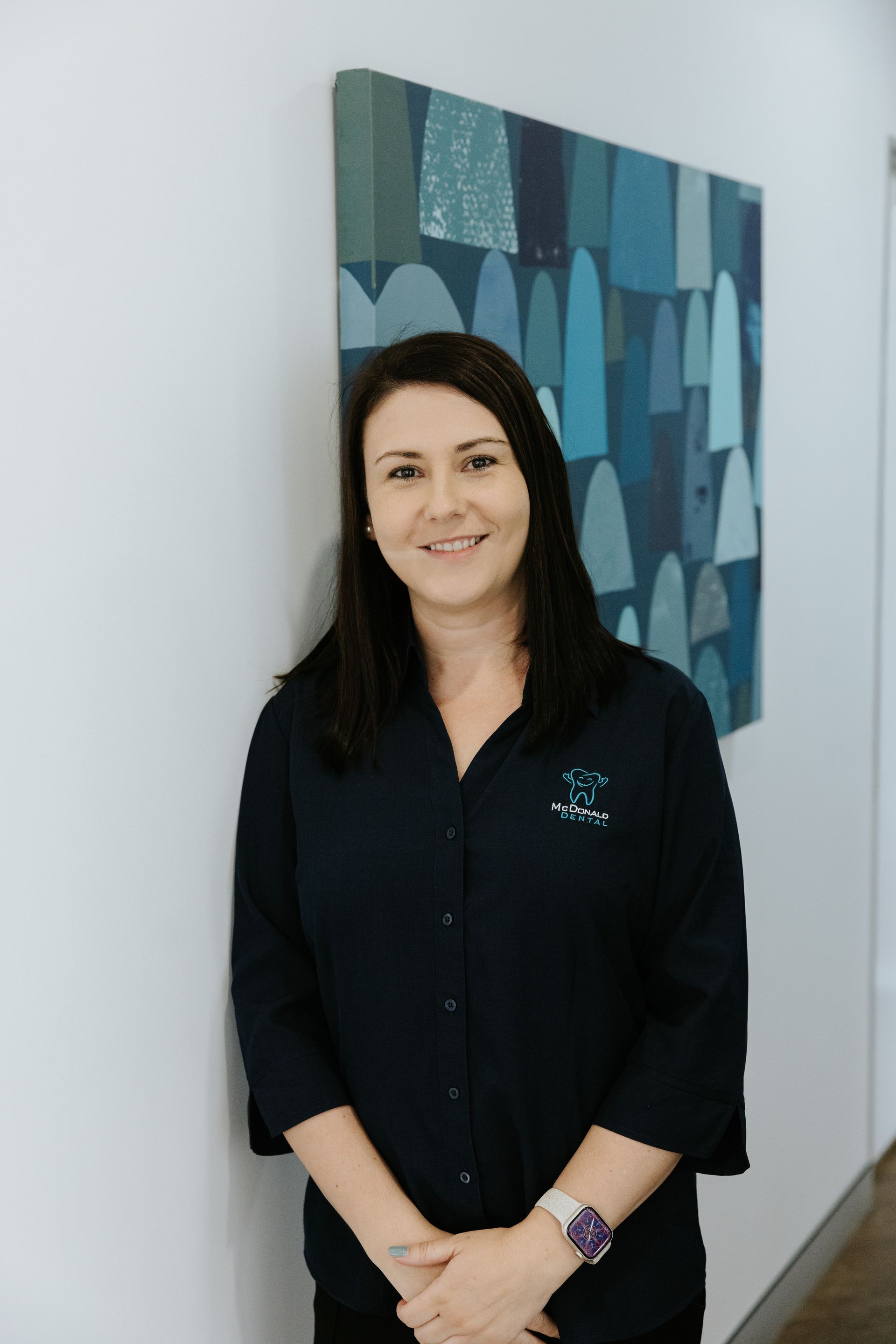 Woman in a blue button-down shirt and black pants, hands in pockets, standing in front of a white background. — McDonald Dental in Gympie, QLD