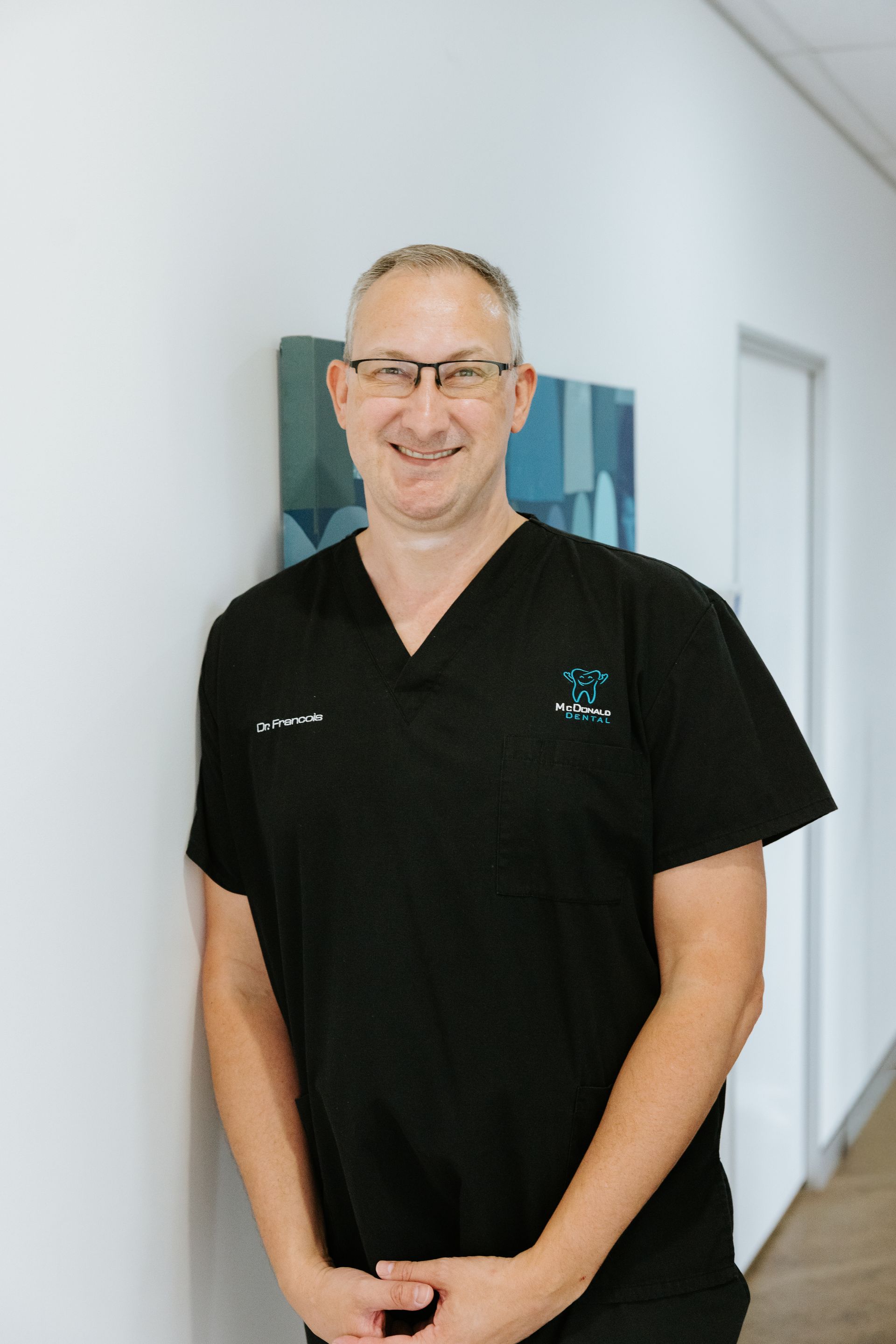 Man in black scrubs and glasses, hands in pockets, smiling against a white background. — McDonald Dental in Gympie, QLD