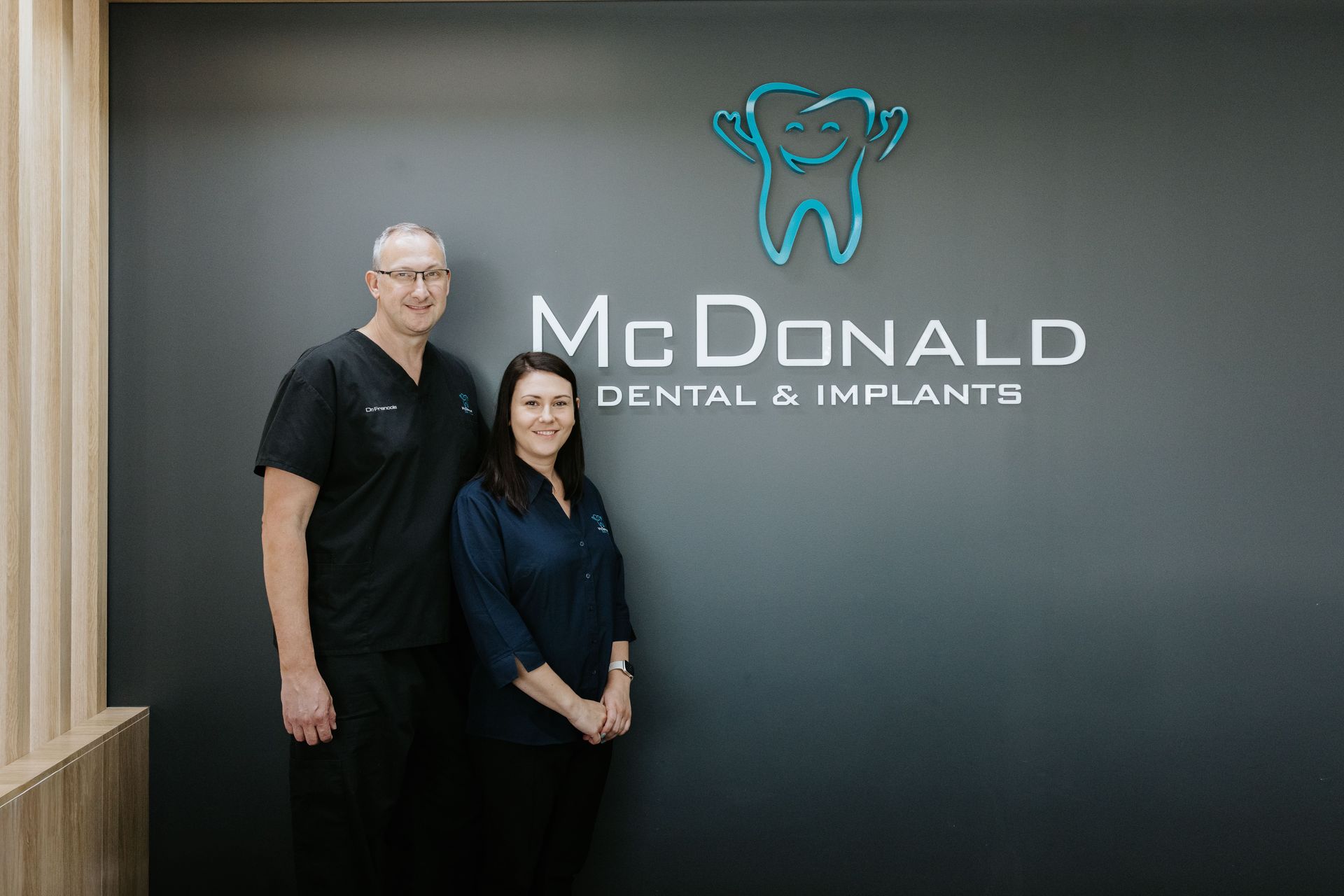 Two people, a man and a woman, stand next to a sign for McDonald Dental & Implants with a tooth logo. — McDonald Dental in Gympie, QLD
