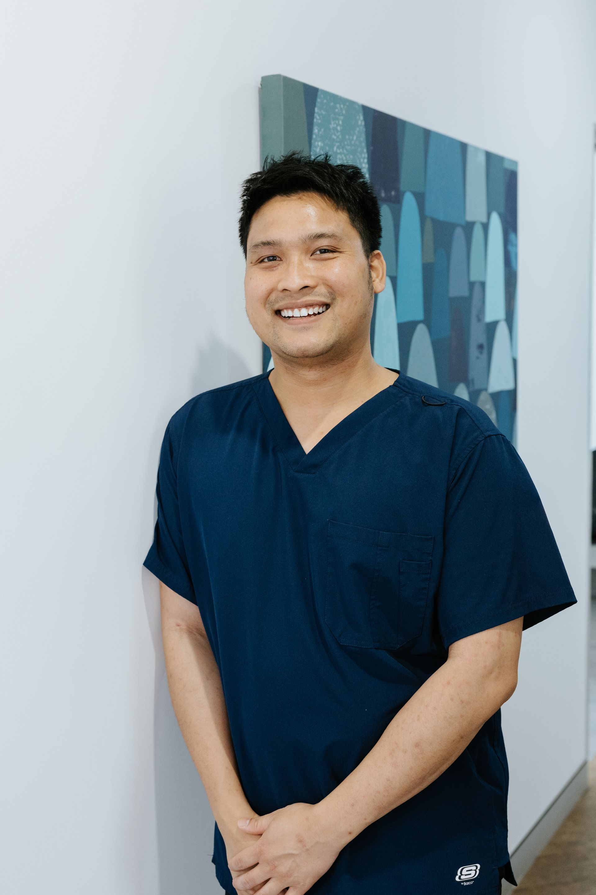 Man in blue scrubs and glasses smiling. Hands in pockets. White background. — McDonald Dental in Gympie, QLD