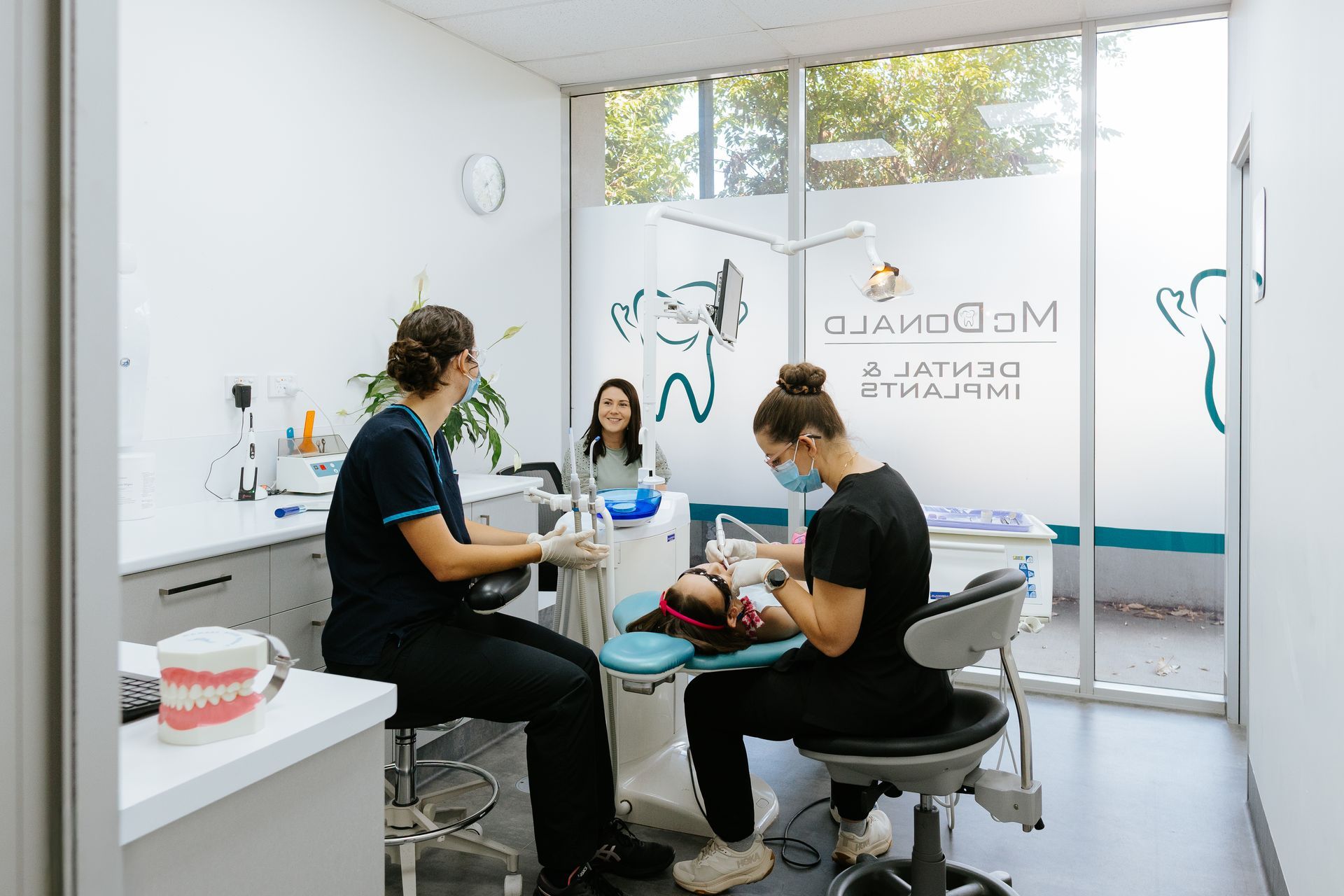 Two dental hygienists treating a patient in a bright dental office; a model of teeth sits on the counter. — McDonald Dental in Gympie, QLD