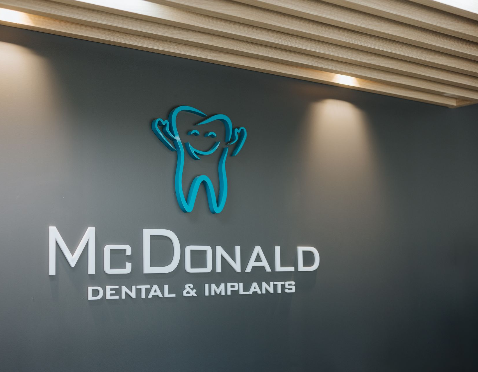 McDonald Dental & Implants logo with smiling tooth icon, teal outline, on gray wall. — McDonald Dental in Gympie, QLD
